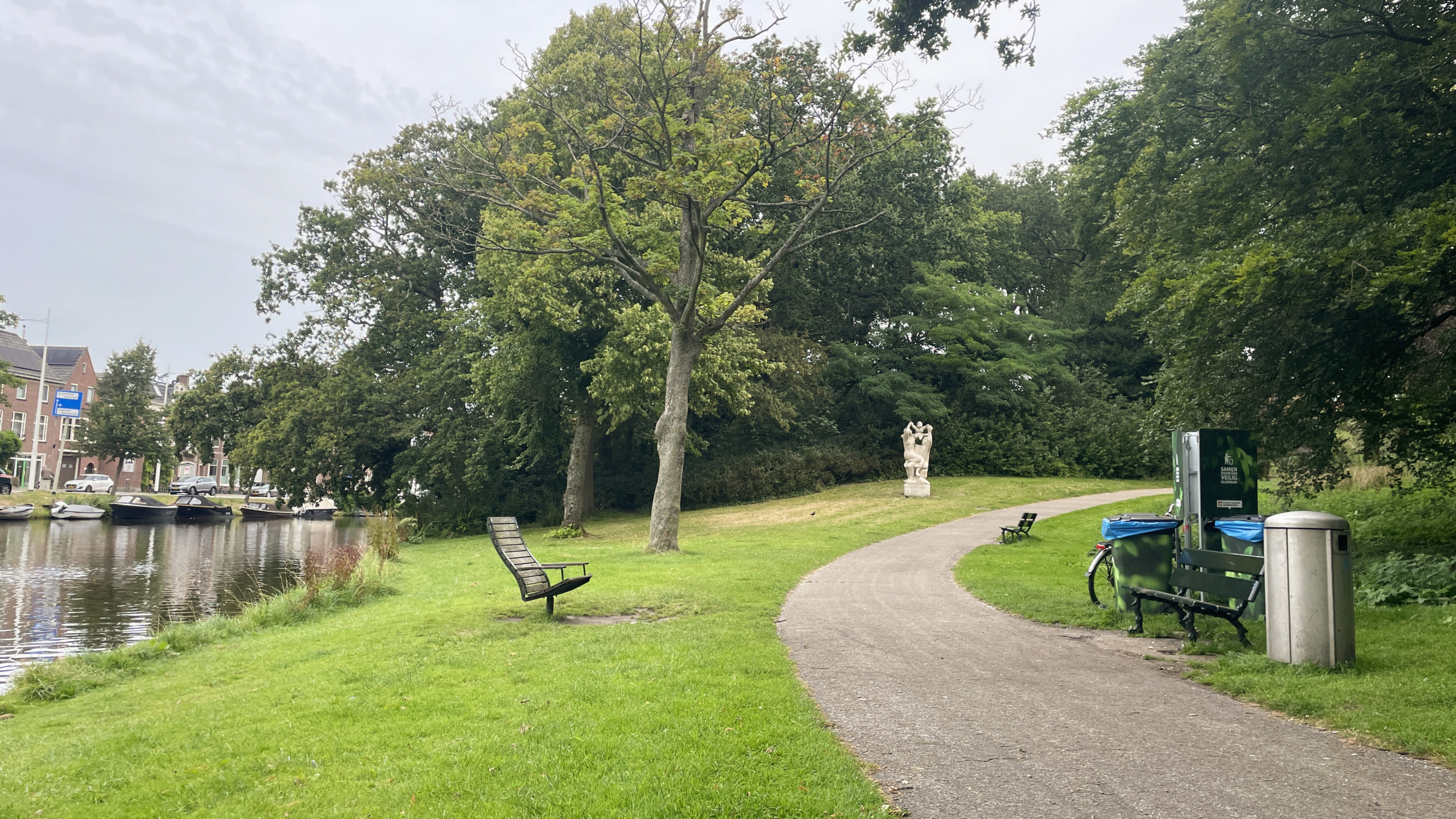 Park met wandelpad, bankjes, beeldhouwwerk, bomen en een waterkanaal met aangemeerde boten.