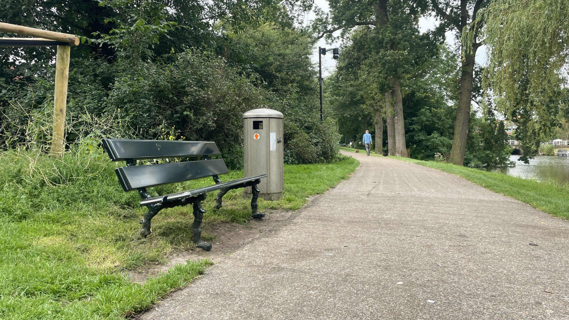 Bankje en afvalbak langs een wandelpad in een park, met een wandelaar op de achtergrond.