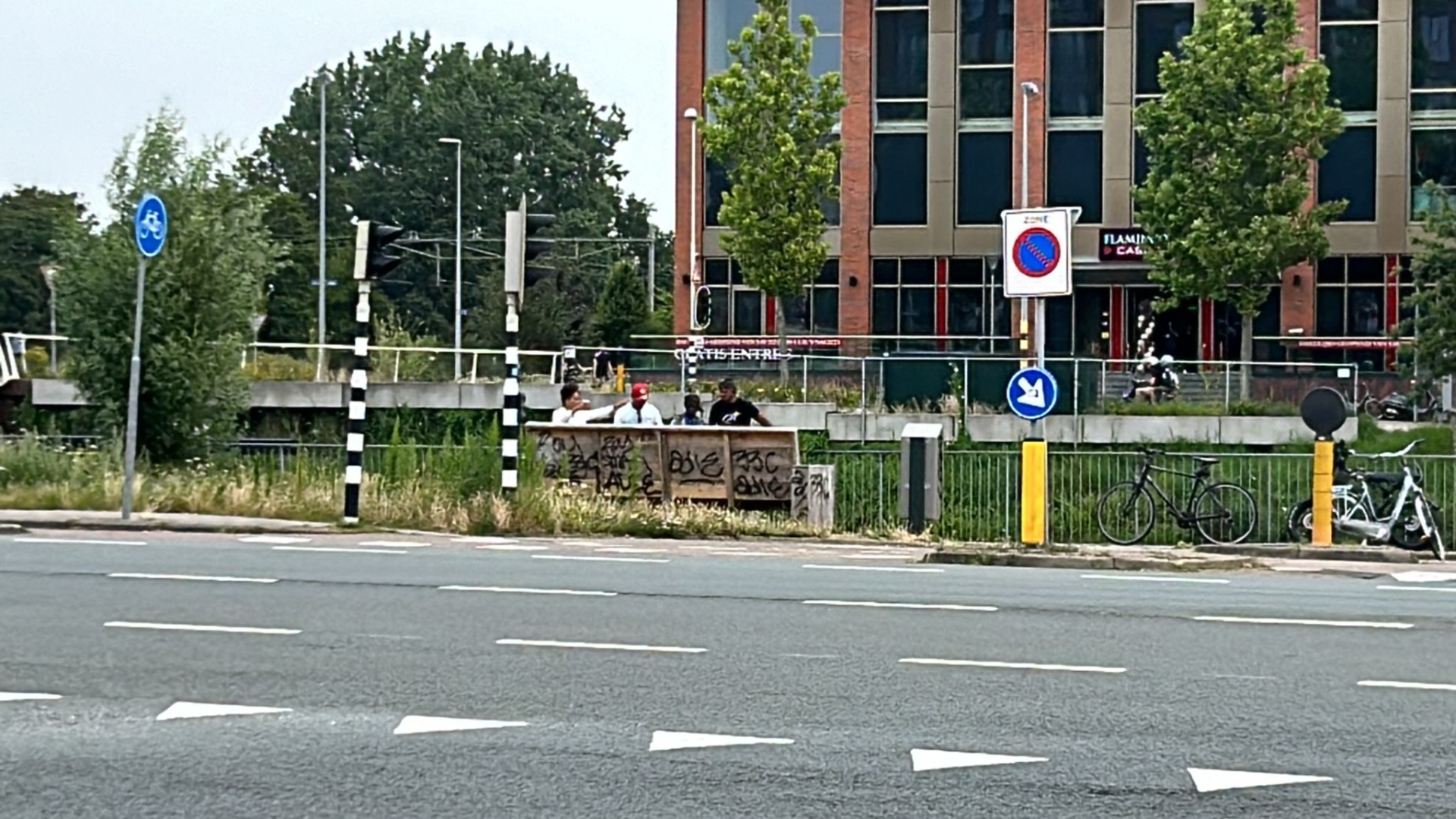 Vier mensen staan bij een met graffiti bedekt gebouw langs een weg met verkeersborden en fietsen, met een modern gebouw op de achtergrond.