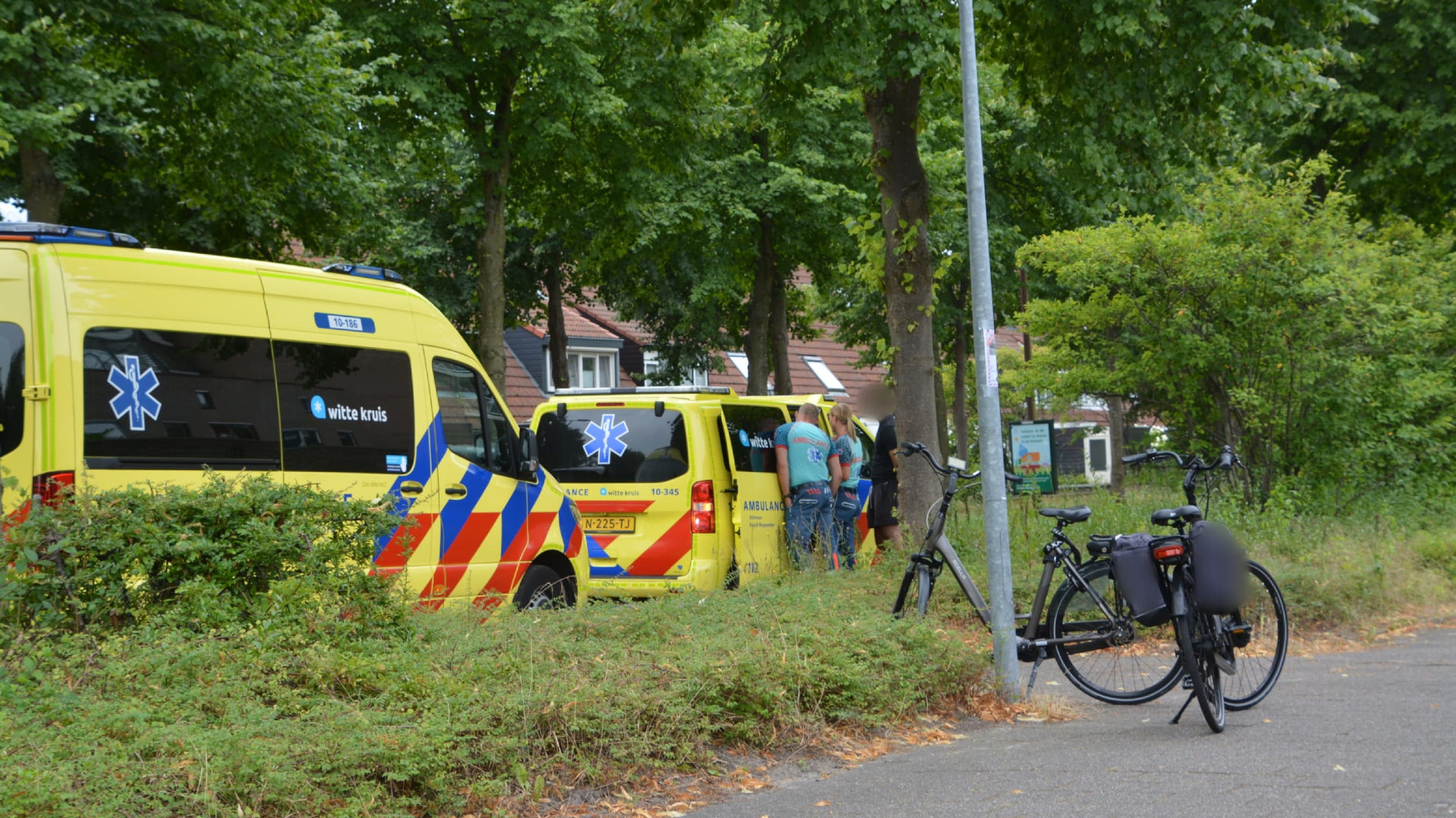 Een straat met twee geparkeerde ambulances en mensen die bij de achterkant van een van de ambulances staan, naast een bosrijke omgeving en meerdere fietsen.