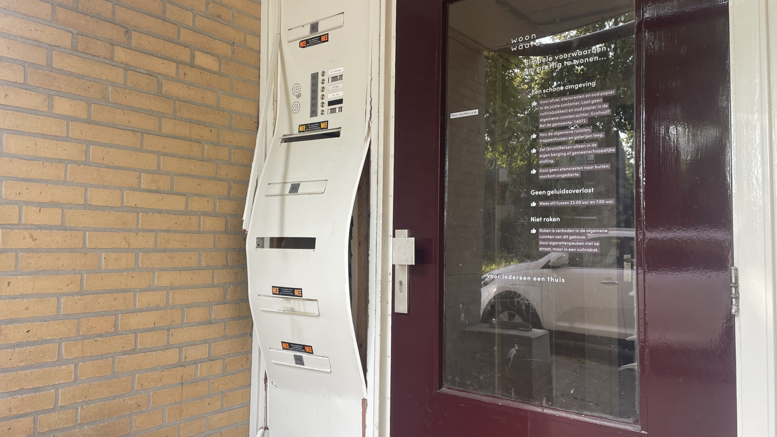 Ingang van een appartementencomplex met een kapotte intercom en brievenbussysteem, naast een deur met regels voor bewoners op een glazen paneel.