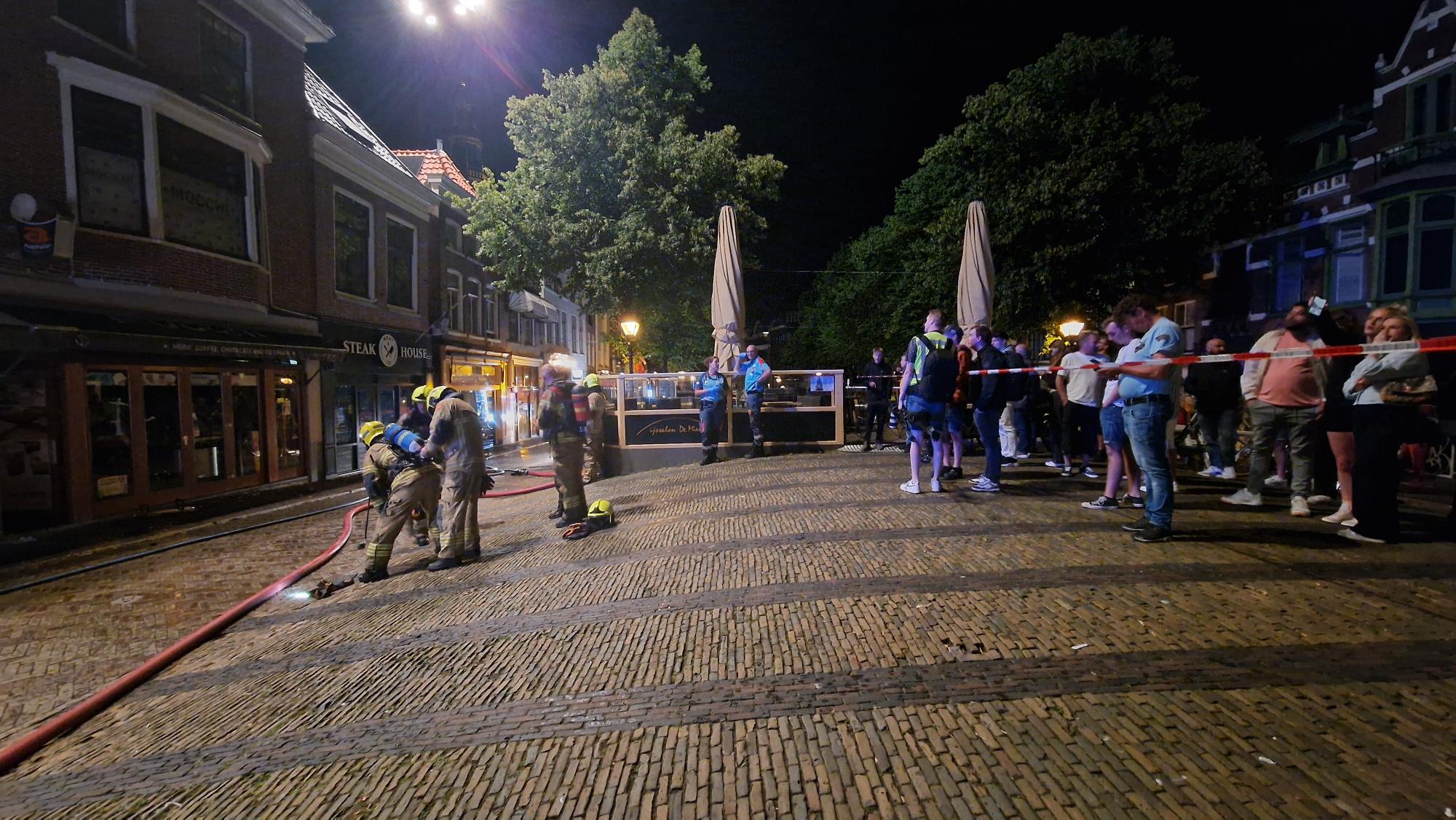 Brandweerlieden werken 's nachts op een geplaveide straat, omgeven door omstanders achter een afzetlint.