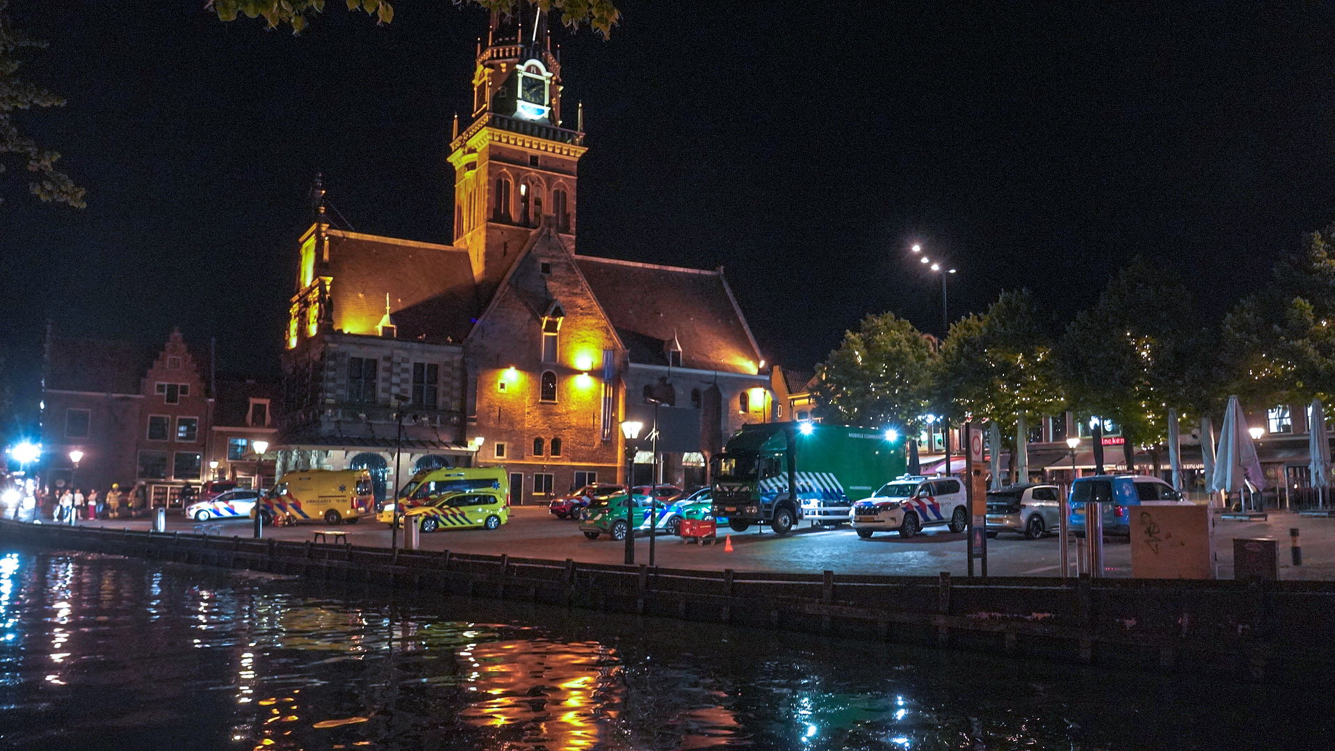 De Waag in Alkmaar 's nachts, omringd door politie- en ambulancevoertuigen, met reflectie in het water.