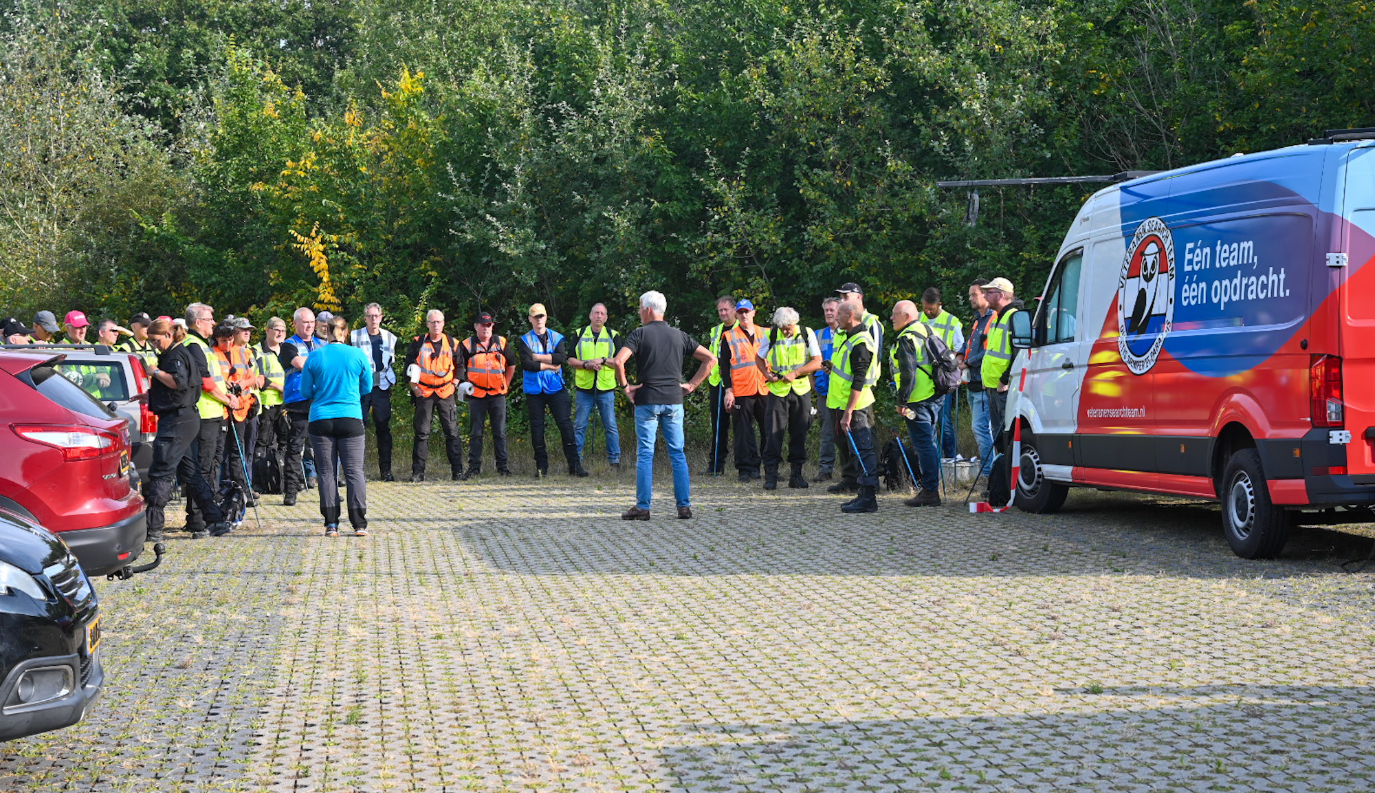 Groep mensen met veiligheidsvesten verzameld op een parkeerterrein bij een witte bestelwagen met de tekst: "Eén team, één opdracht".