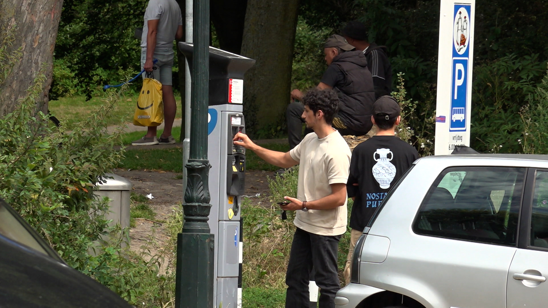 Man bij parkeerautomaat in een park omgeven door bomen en andere mensen op de achtergrond.