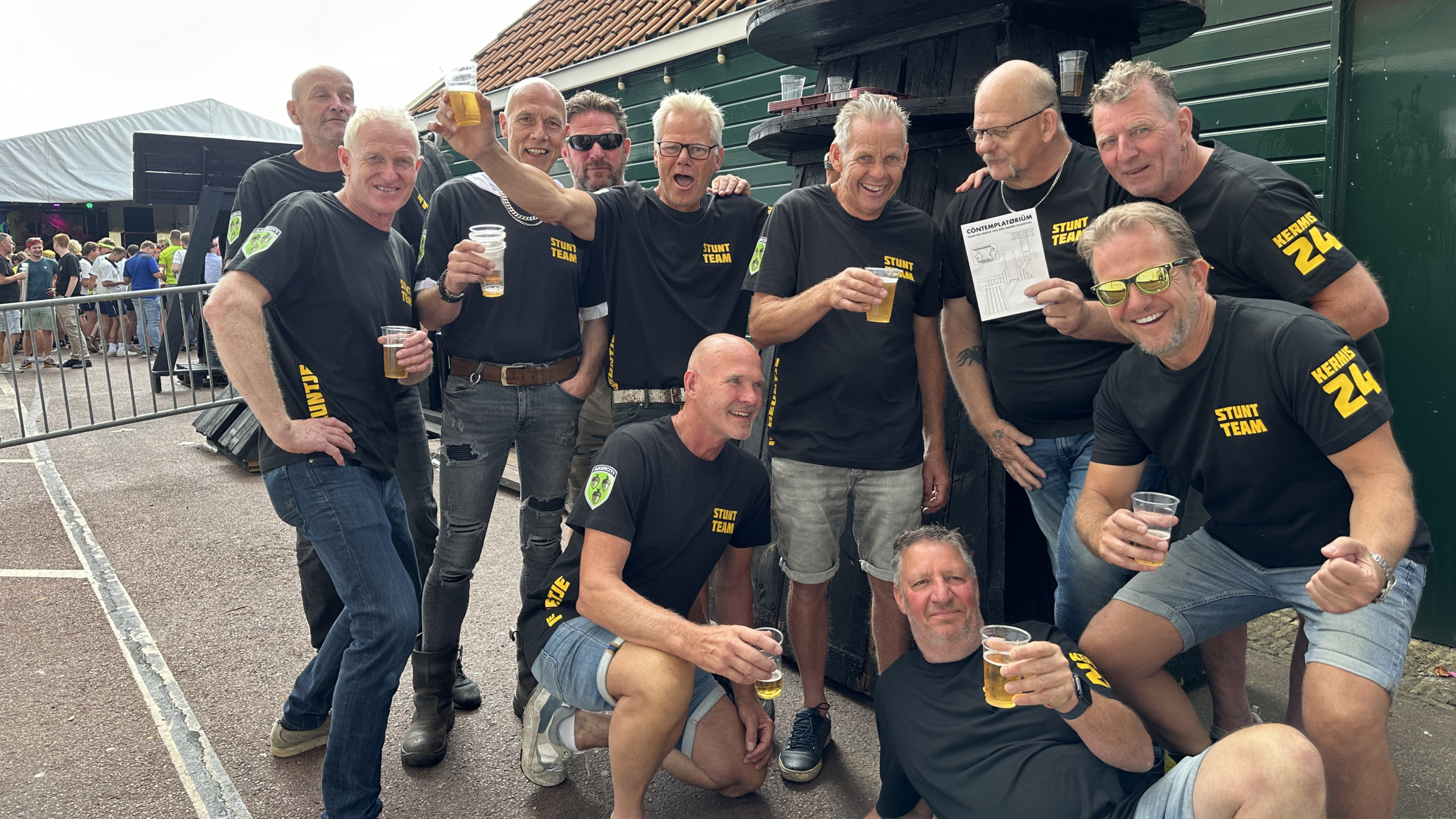 Een groep mannen in zwarte T-shirts met opschrift "Stunt Team" bij een buitenevenement, lachend en bier drinkend. Contemplatorium replica