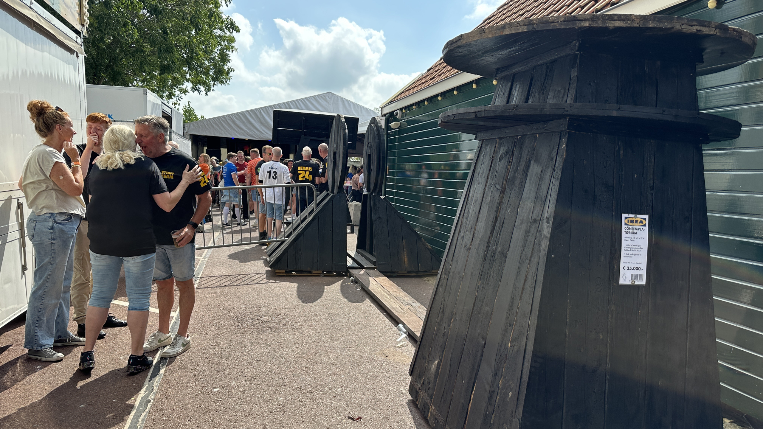 Een groep mensen staat te praten bij een openluchtfestival met kraampjes en een tent. Op de voorgrond staan grote houten kabelrollen met een IKEA-prijskaartje erop aangebracht. Contemplatorium replica