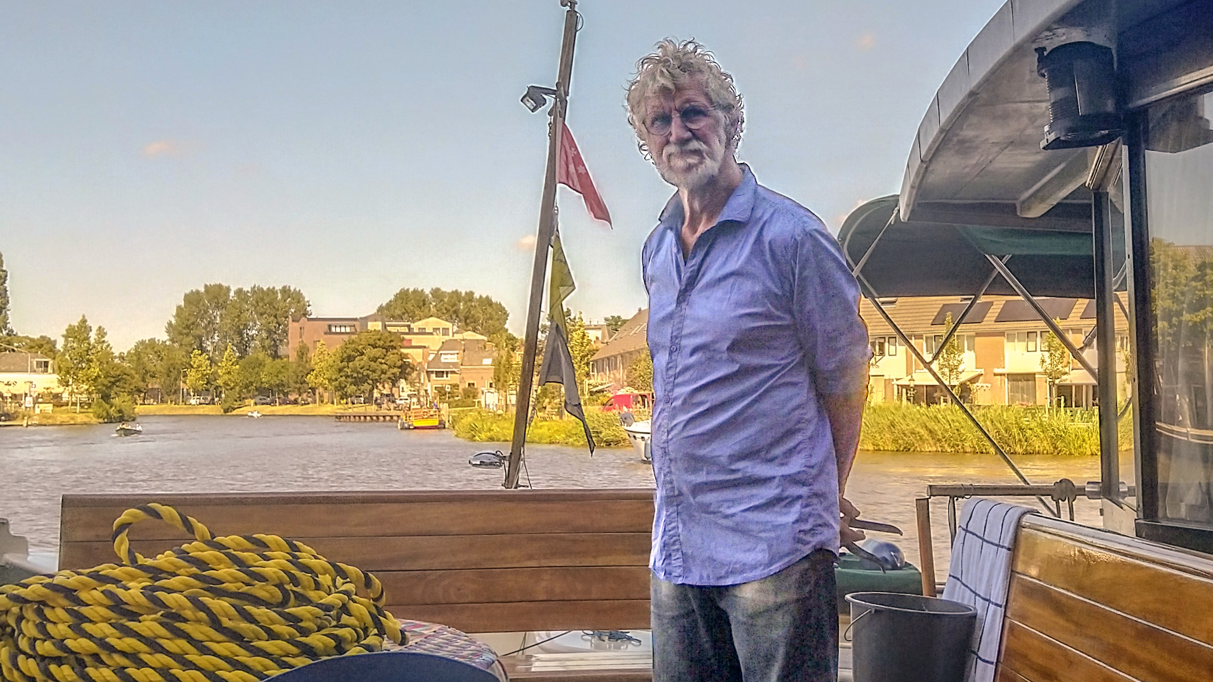 Man op een boot met een blauwe overhemd aan; op de achtergrond een rivier en huizen.