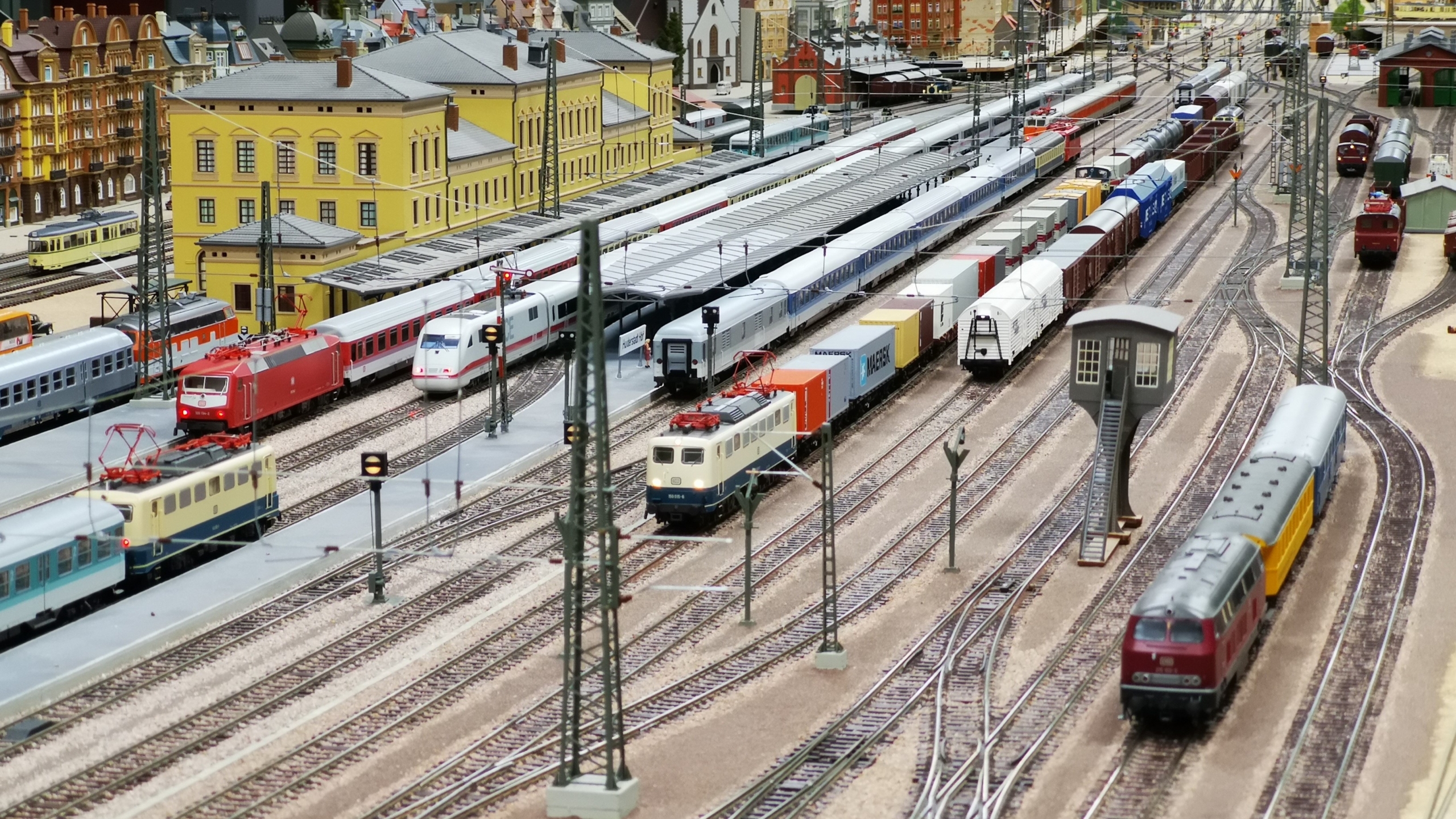 Spoorwegmodel van een druk station met verschillende treinen, wagons en een geel stationsgebouw op de achtergrond.