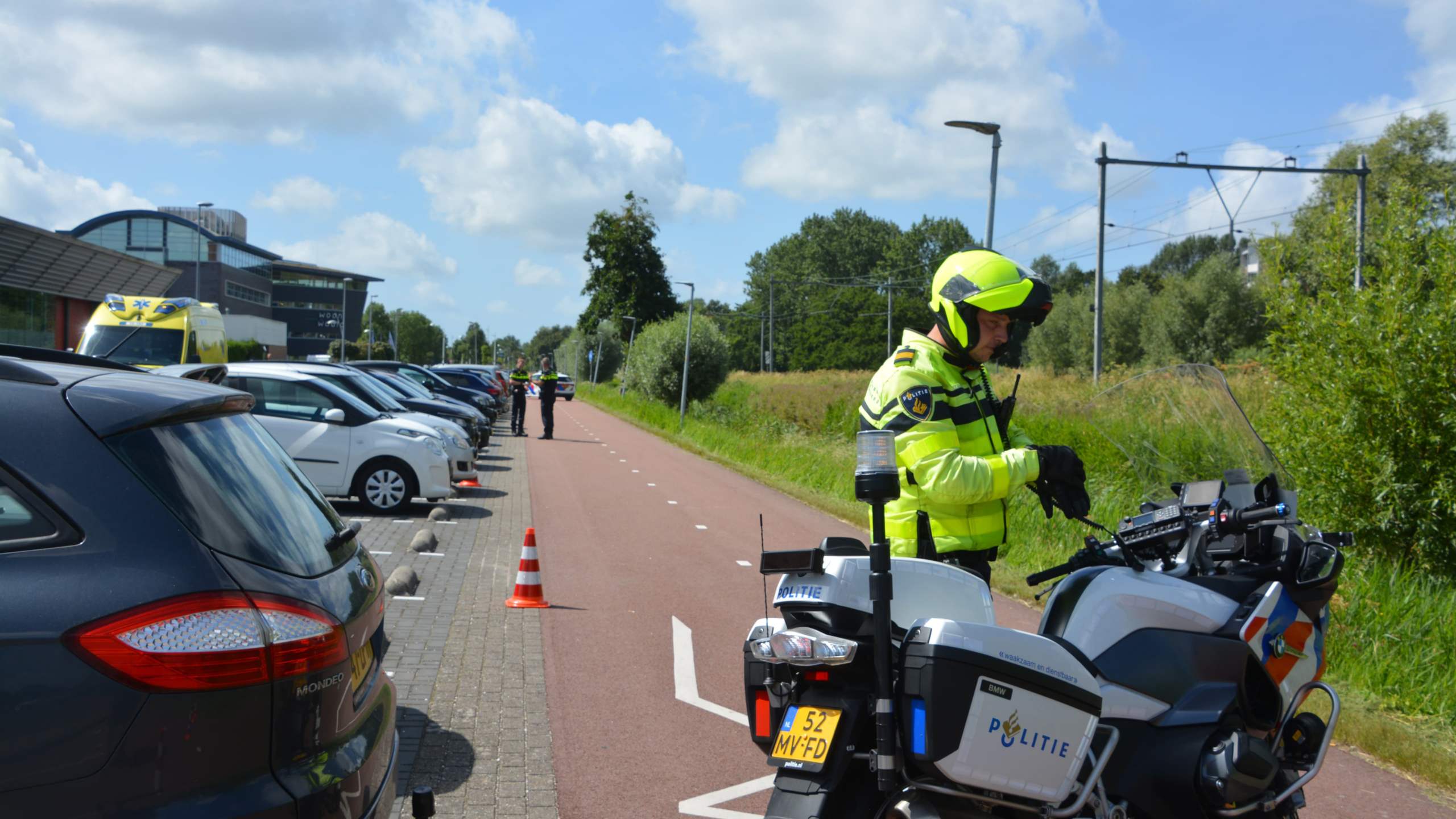 Politiecontrole op een fietspad langs een parkeerplaats met geparkeerde auto's en een geparkeerde ambulance.