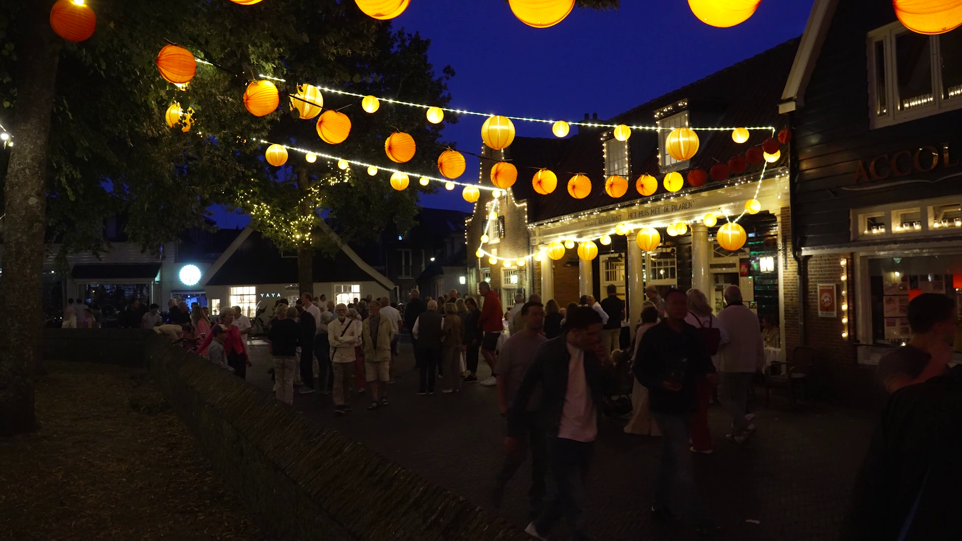 Straatfeest in de avond met mensen onder oranje en gele lantaarns en lichtslingers.