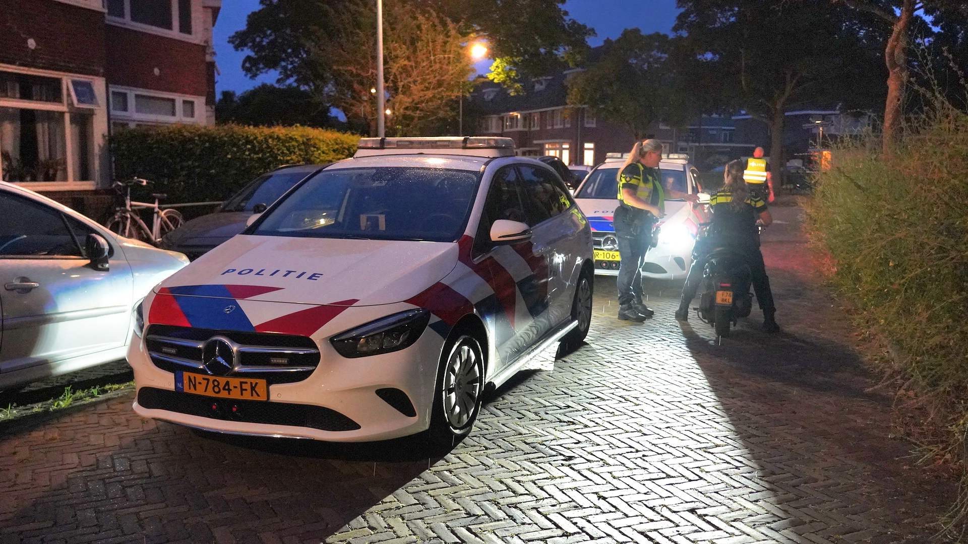 Politieauto's in een woonwijk, twee agenten in gesprek met een persoon op een scooter in de avond.