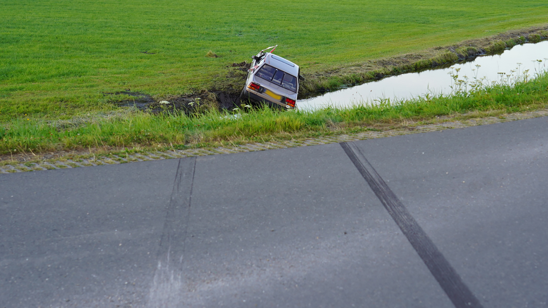 Auto in greppel naast een landweg met bandensporen op het asfalt.