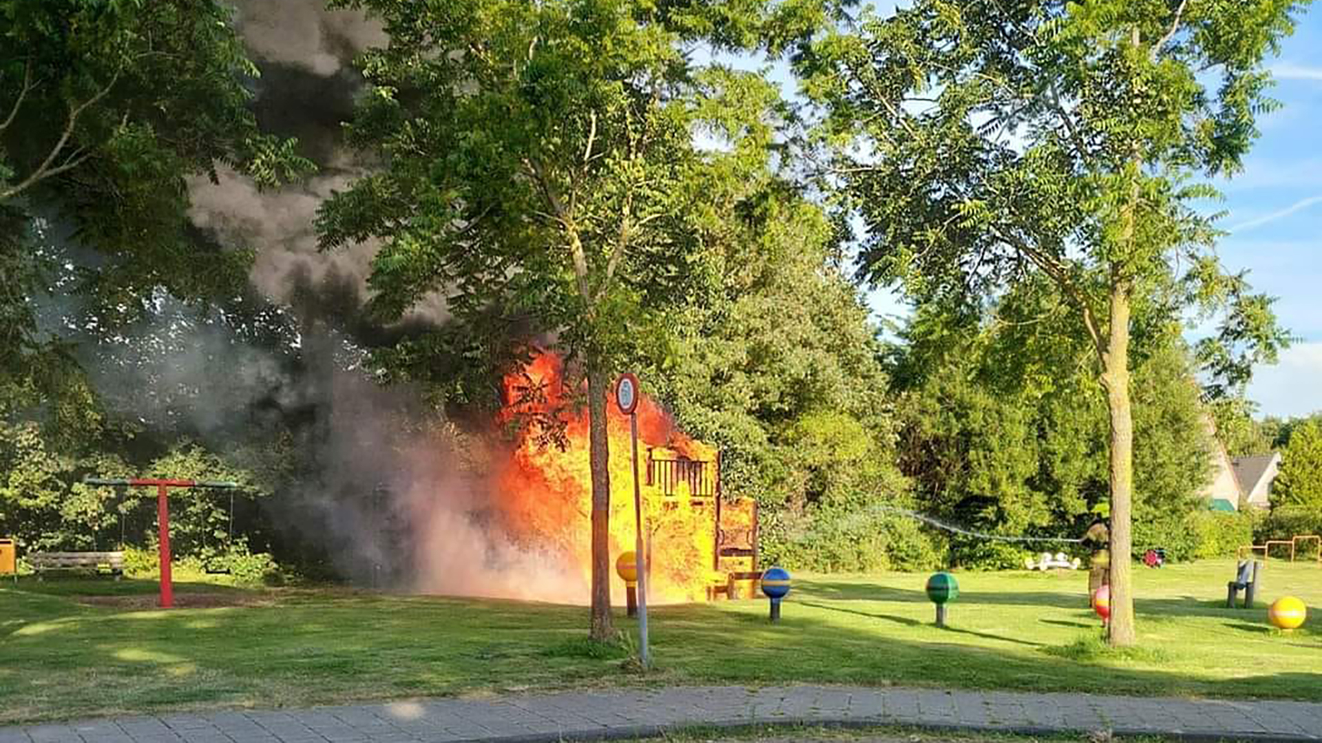 Speeltoestel in een park in brand, met veel rook en vlammen zichtbaar tussen de bomen.