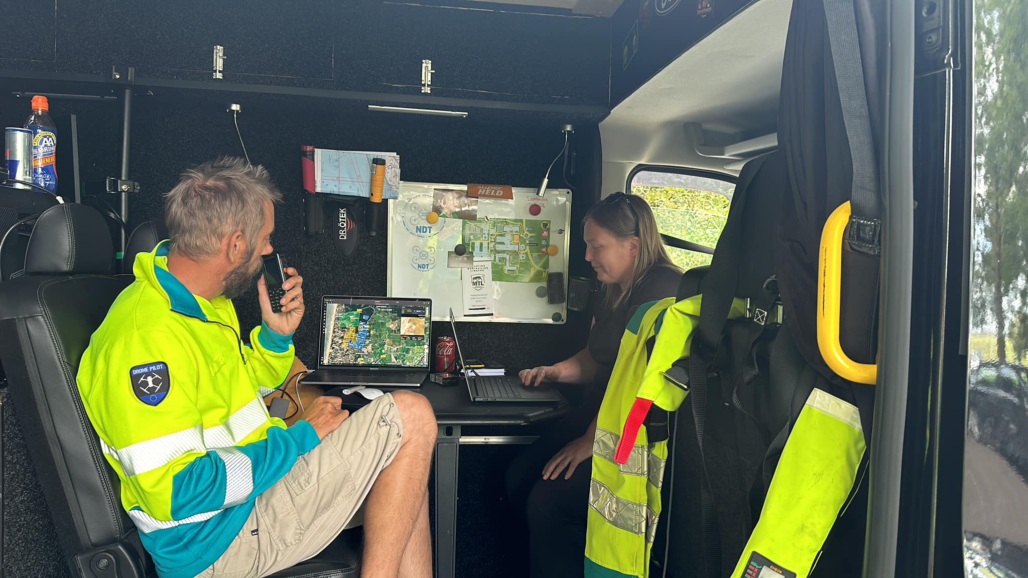 Twee mensen in een mobiele commandopost werken samen; de ene praat in een portofoon, de andere typt op een laptop.