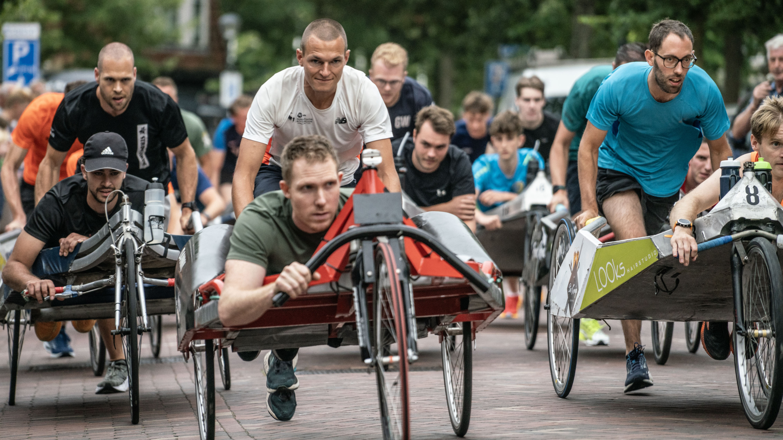 Mensen nemen deel aan een beddenrace met aangepaste transportmiddelen in een stedelijke omgeving, waarbij toeschouwers op de achtergrond zichtbaar zijn.