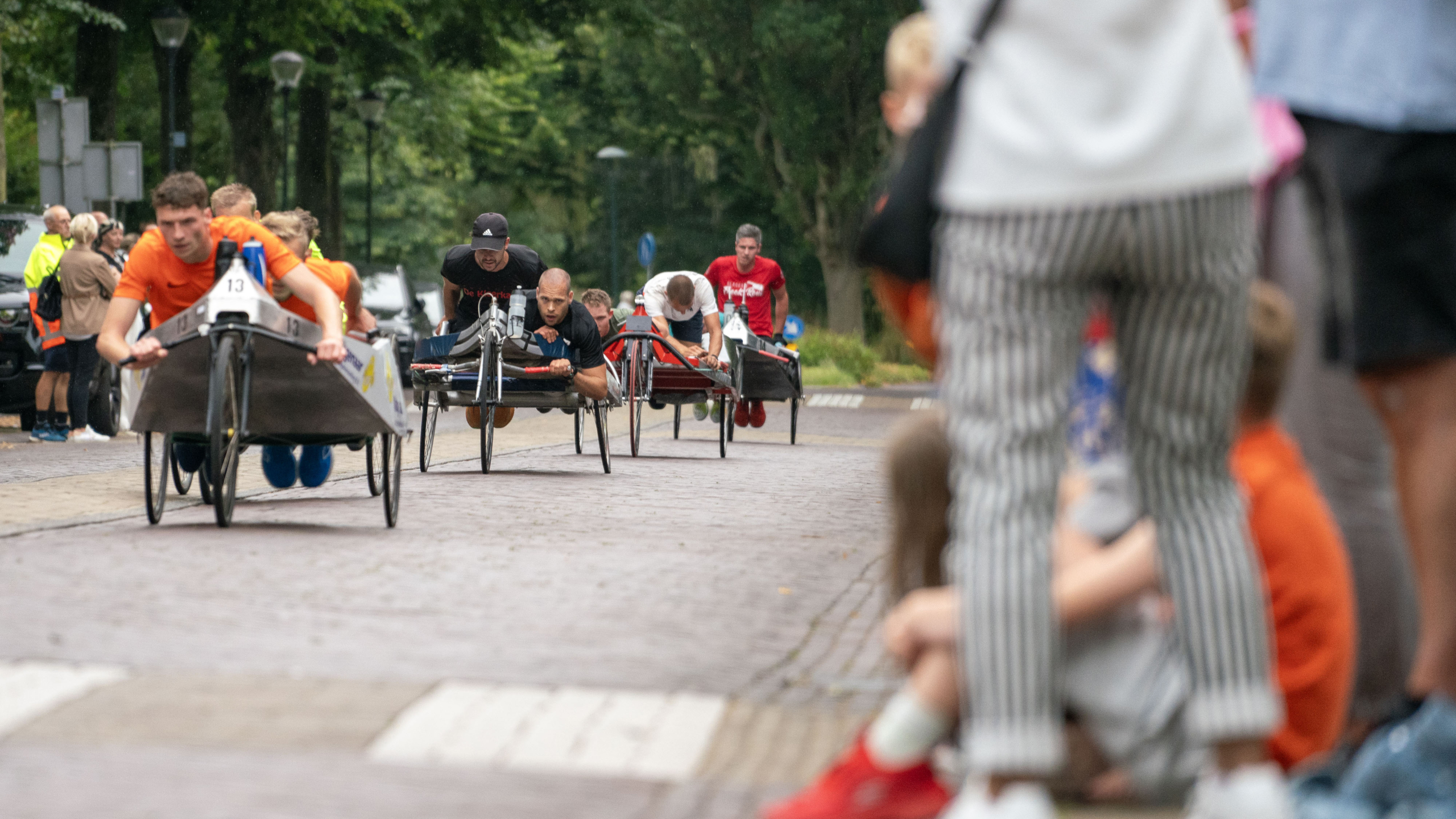 Mensen die deelnemen aan een beddenrace op een geplaveide weg met toeschouwers langs de zijlijn.