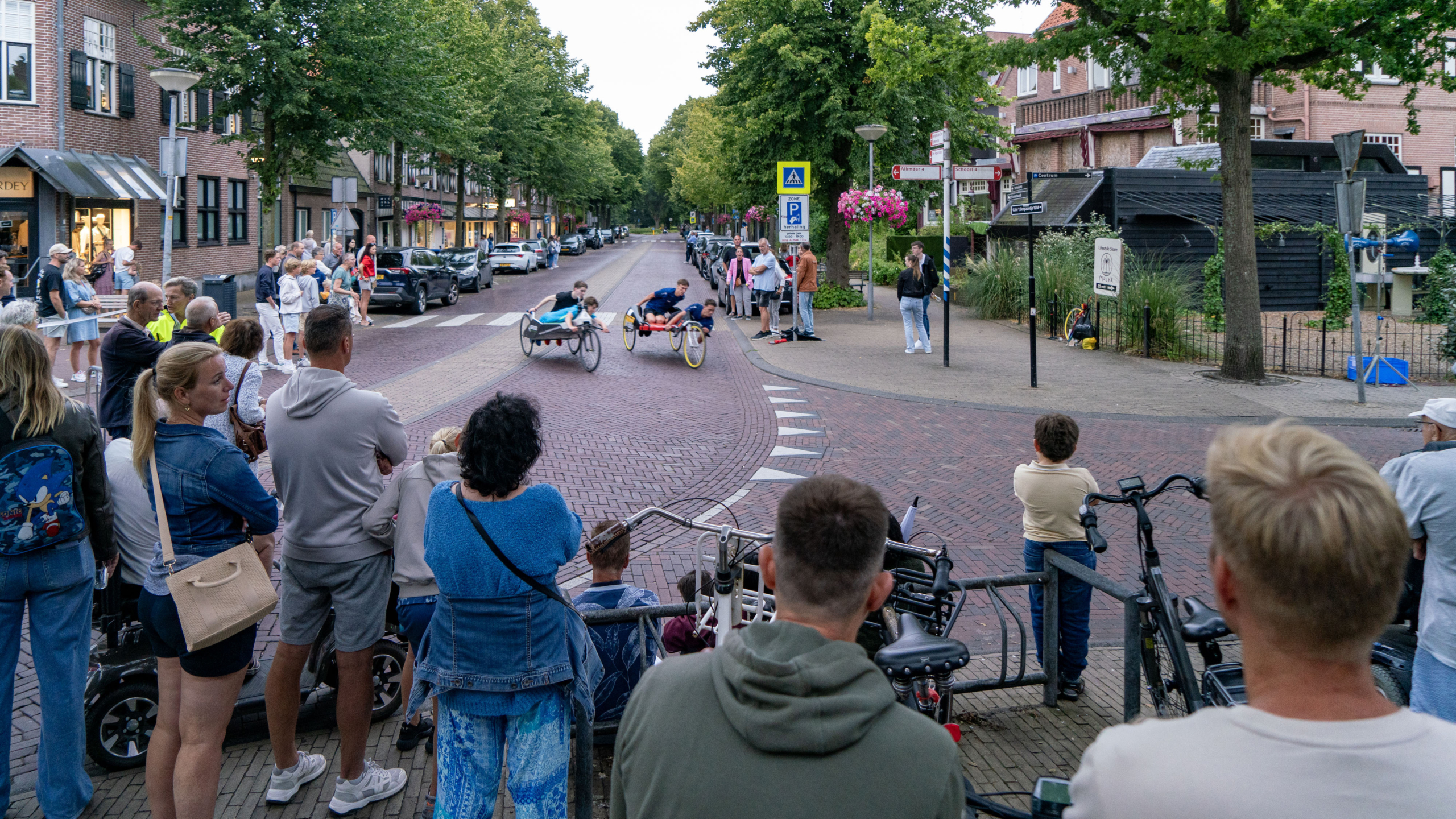 Toeschouwers kijken naar een beddenrace in een straat met bomen en huizen op de achtergrond.