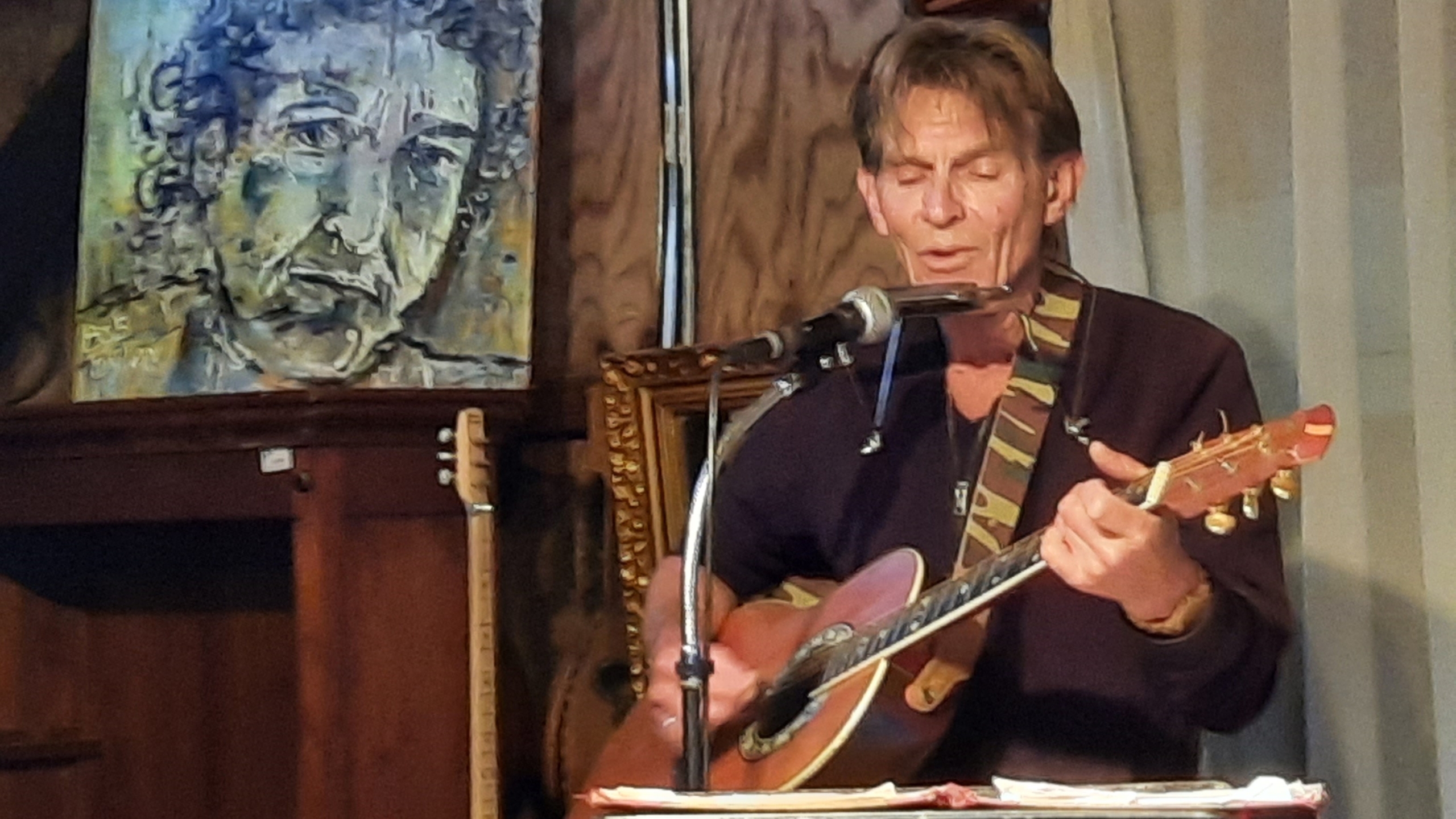 Man speelt gitaar en mondharmonica in een kamer met een schilderij op de achtergrond.