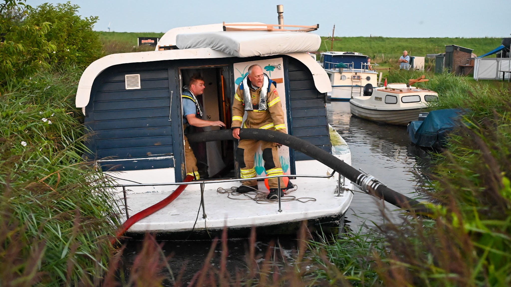 Brandweerlieden in een woonboot aan het werk met een grote slang, omringd door water en riet.