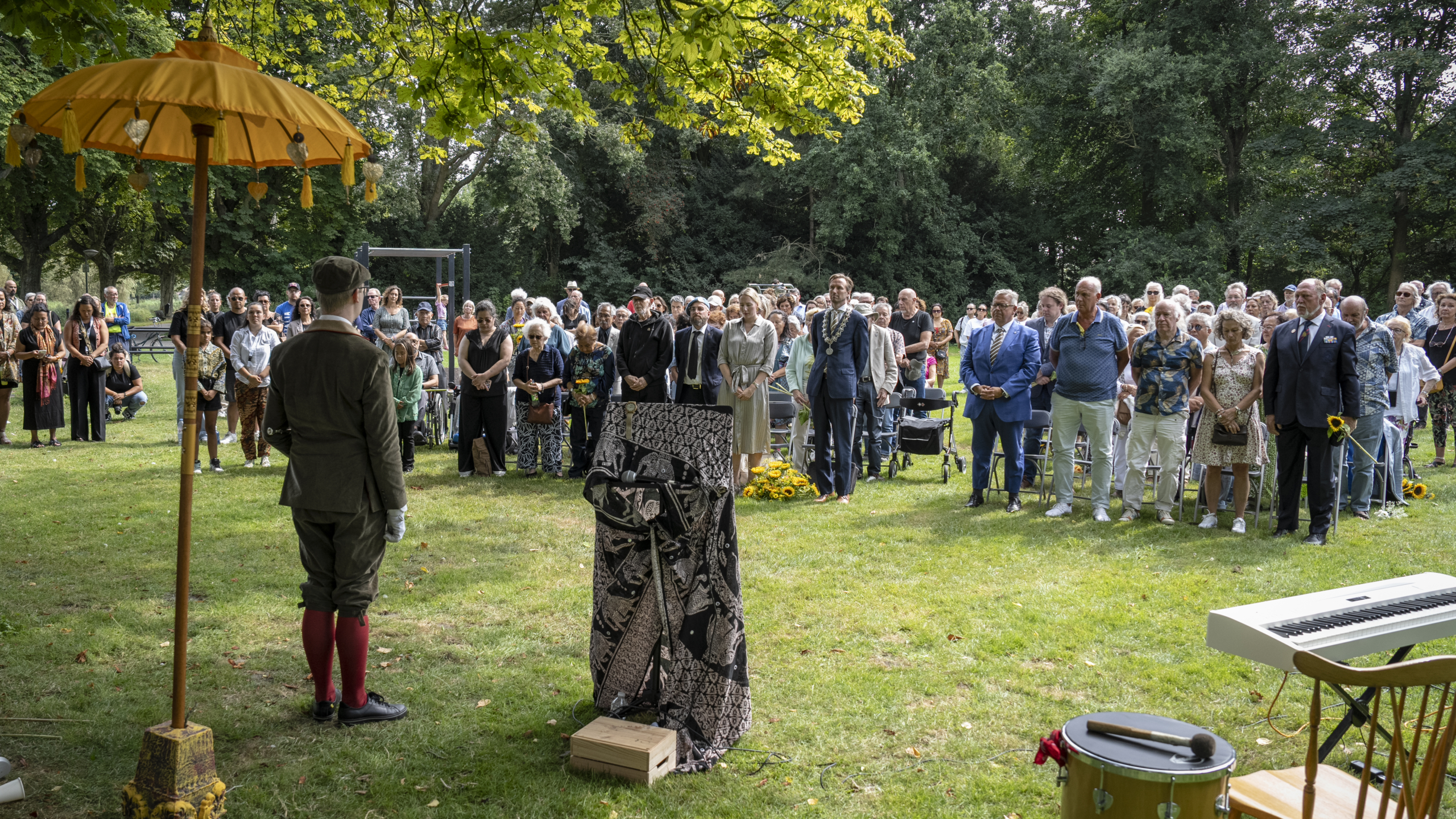 Mensen staan samen in een park tijdens een ceremonie onder een gele parasol.