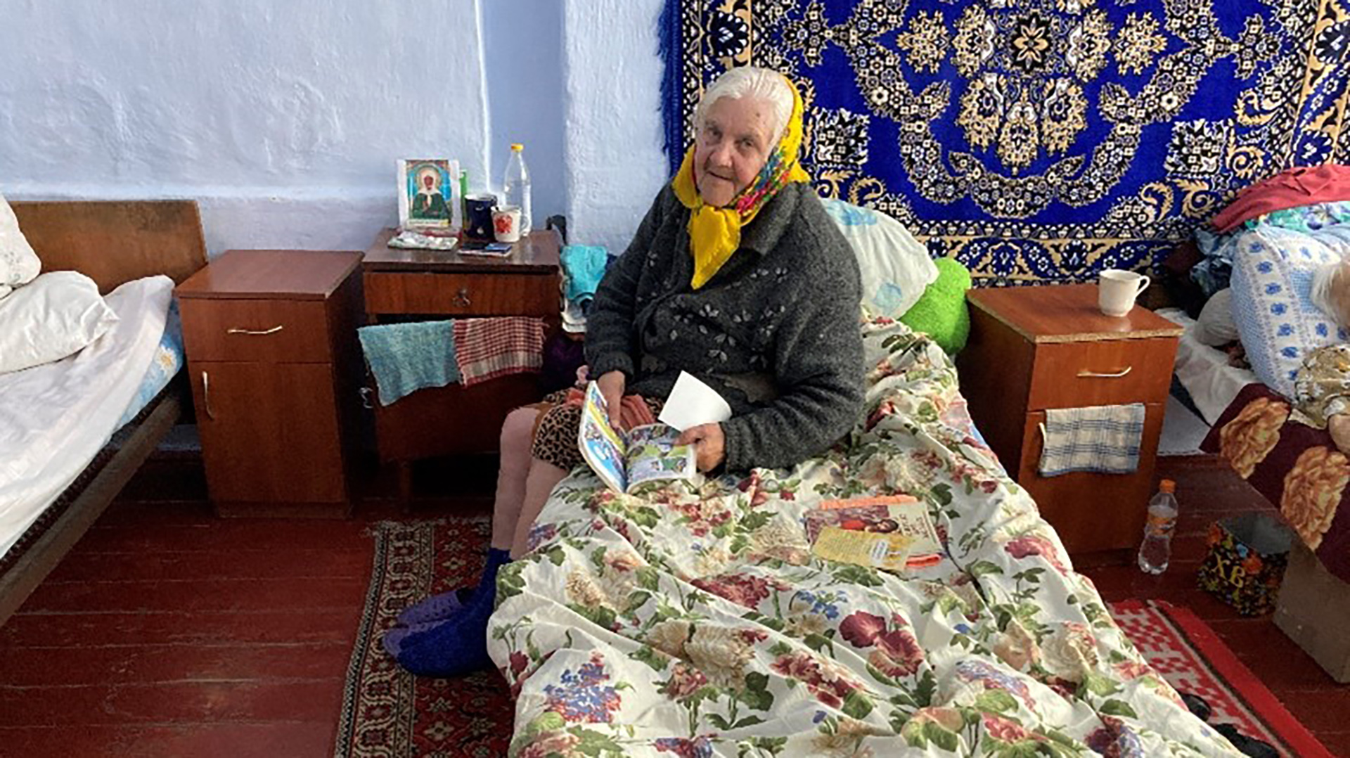 Oudere vrouw zittend op een bed in een eenvoudige kamer, lezend uit een tijdschrift, met kleurrijk tapijt aan de muur.