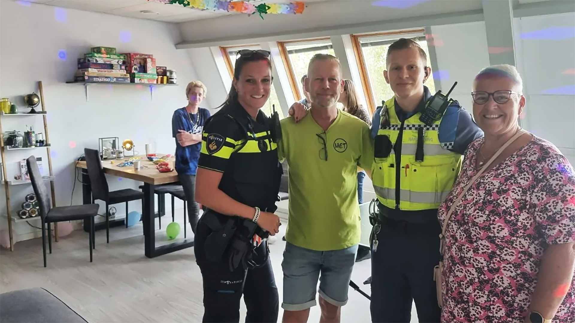 Vier mensen poseren voor de camera, waarvan twee in politie-uniform, in een kamer met een tafel en spelletjes op een plank.