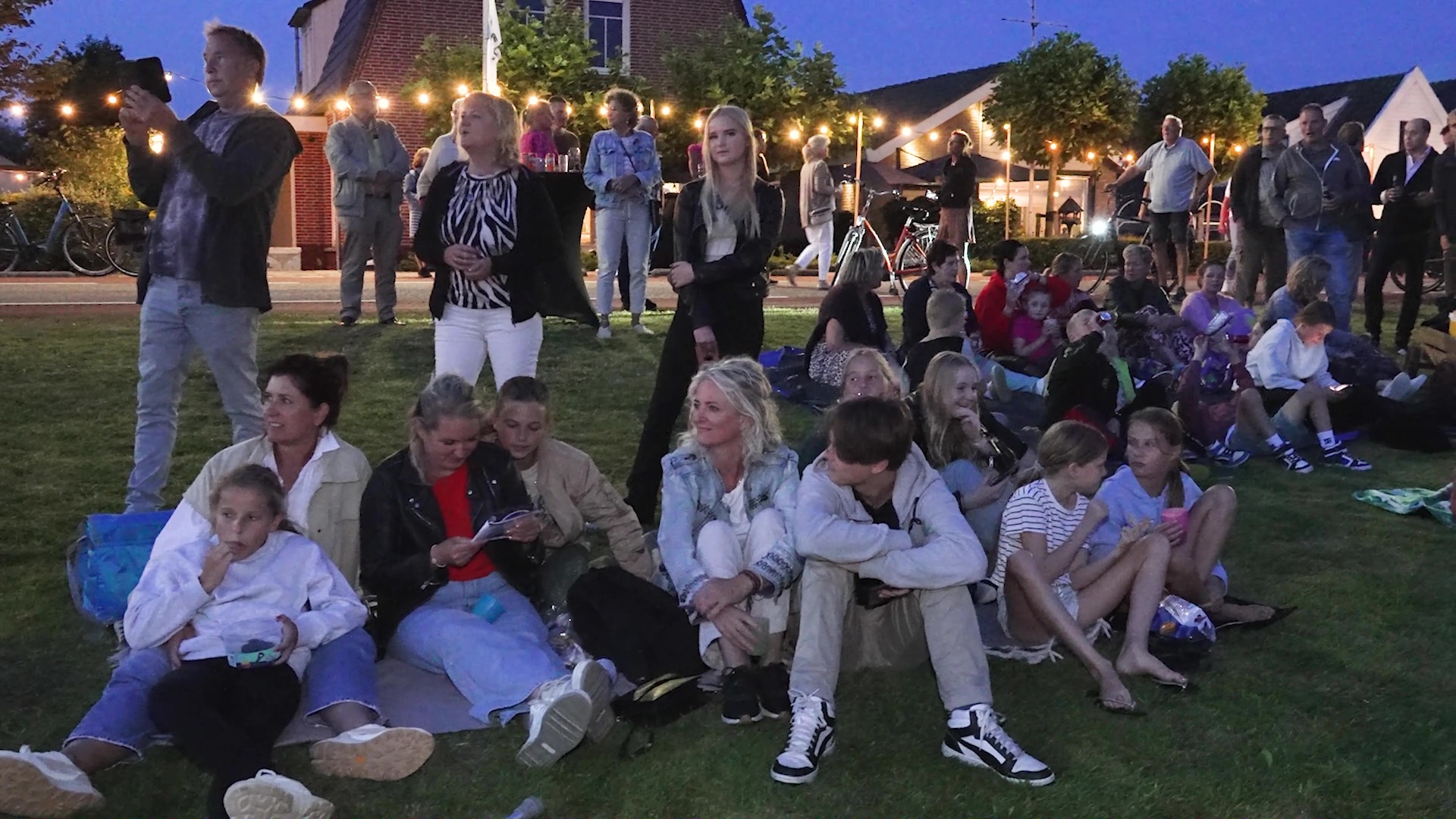 Groep mensen geniet van een avond buiten op gras, sommige zittend op dekens en anderen staand, met sfeerverlichting op de achtergrond.