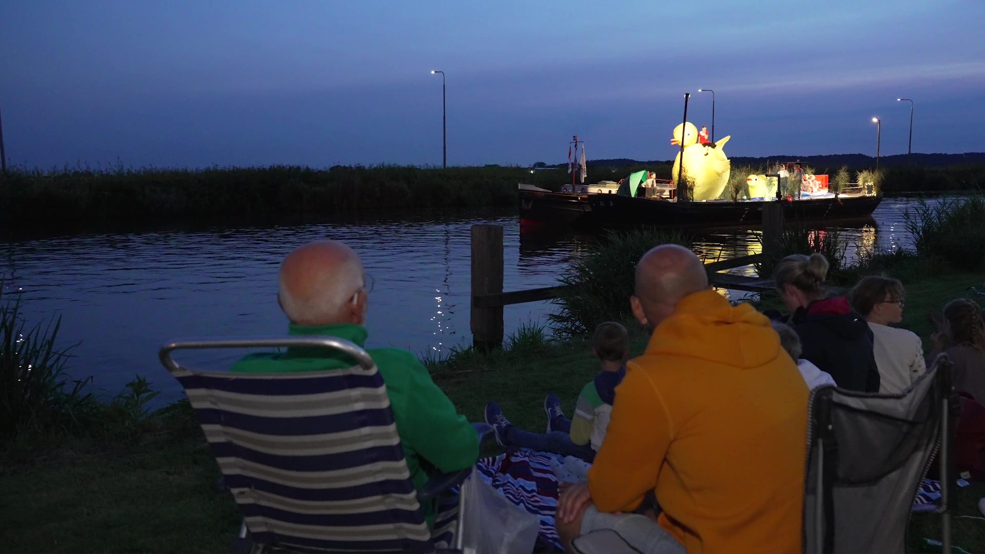 Mensen kijken 's avonds naar een verlichte paradeboot met een grote opblaasbare eend in het water.