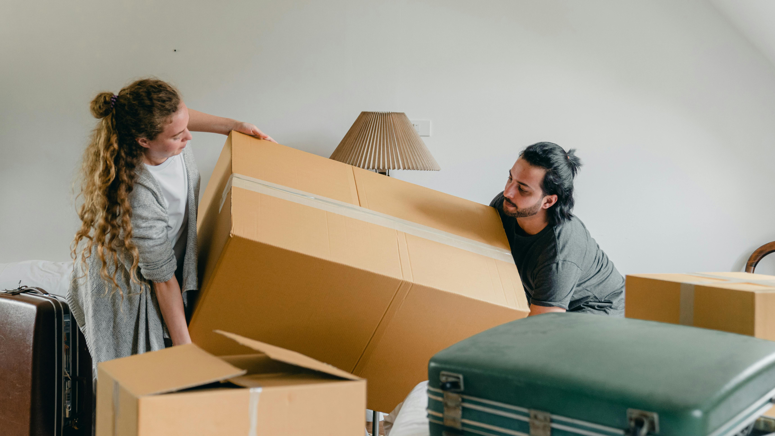 Man en vrouw dragen grote kartonnen doos, omringd door andere verhuisdozen en bagage.