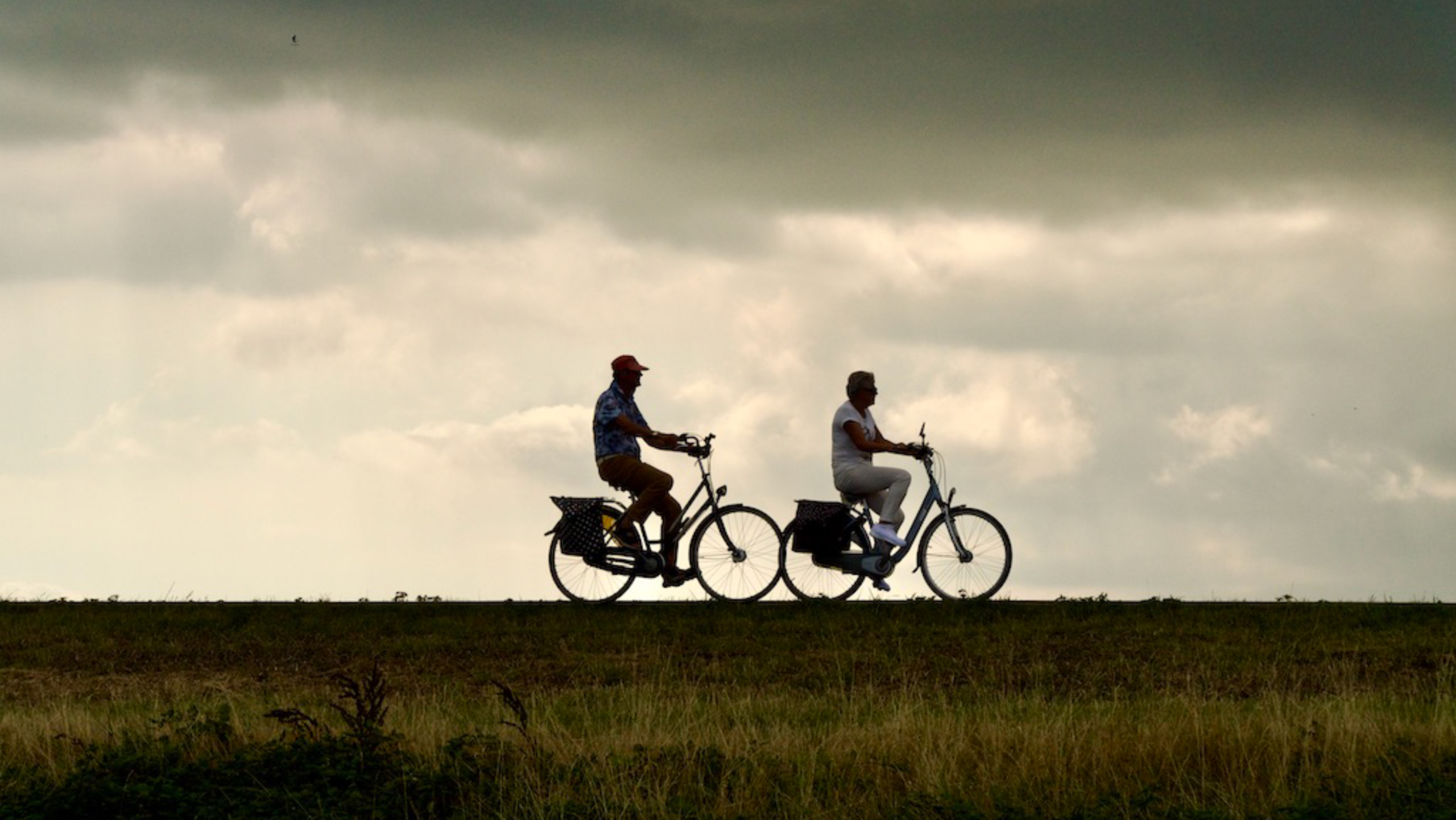 Twee mensen fietsen op een dijk tegen een bewolkte hemel.