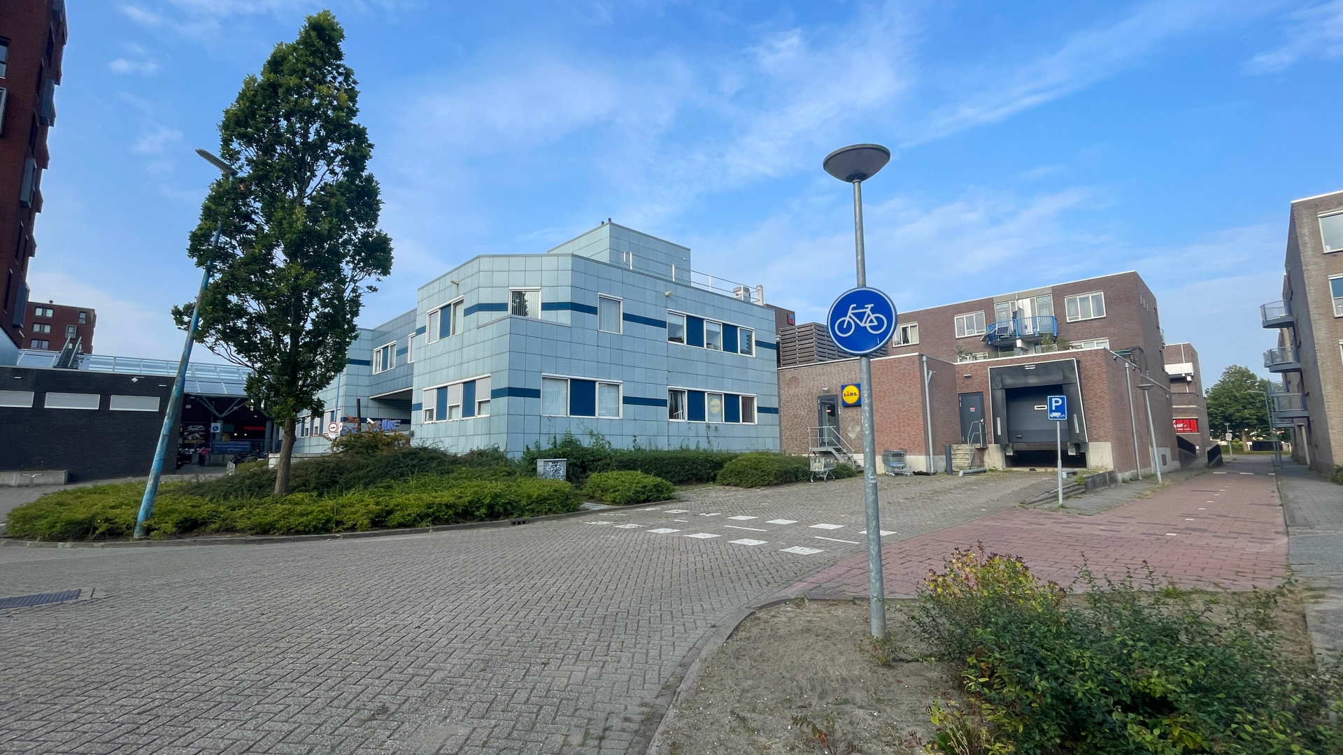 Fietspad teken bij blauw-grijs gebouw, met parkeerplaats en Lidl supermarkt op de achtergrond.