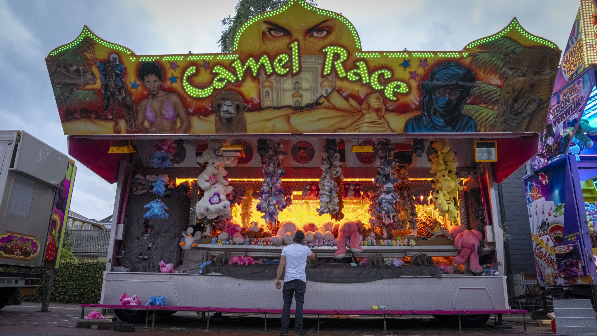 Kermisgames kraam met lichtgevende "Camel Race" bord en diverse kleurrijke pluche prijzen, met een man in witte T-shirt ervoor.