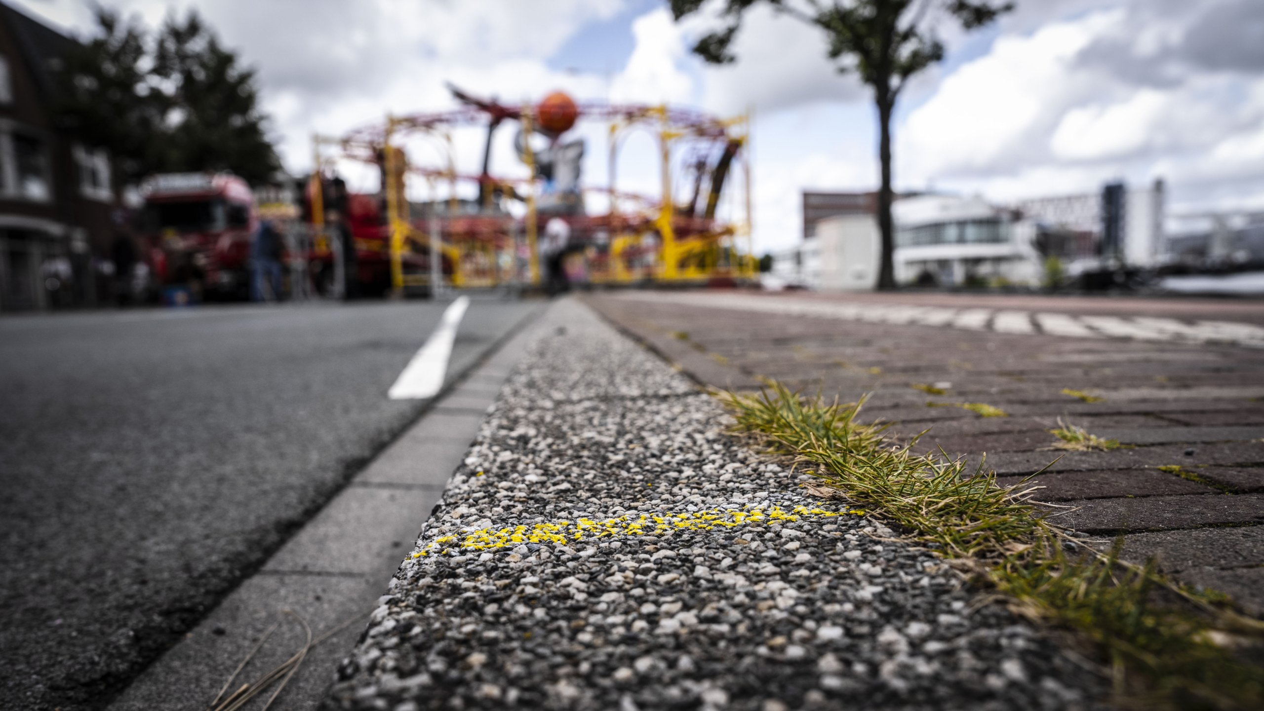 Close-up van een trottoir met een rand van grind en een gele streep, met onscherpe achtergrond van gebouwen en een kermisattractie.