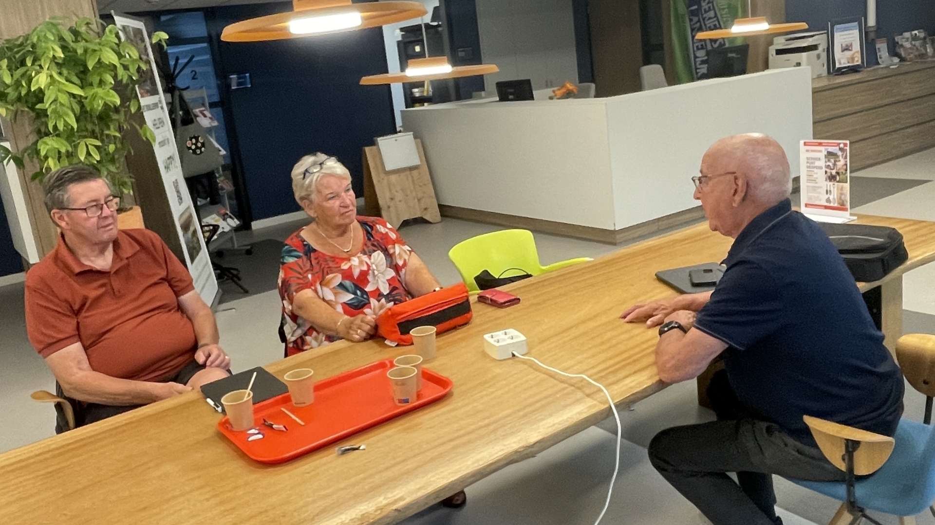 Drie oudere mensen zitten aan een tafel in een moderne ruimte, in gesprek met elkaar.