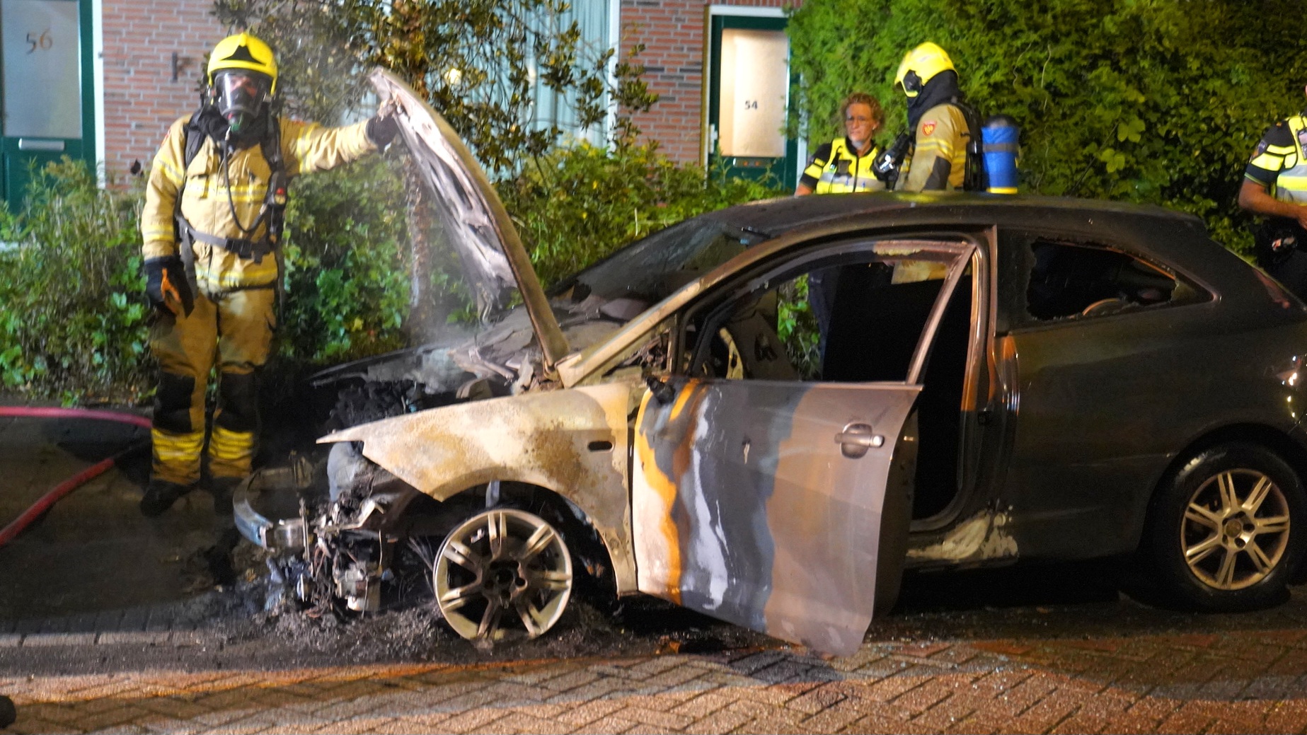 Brandweermannen blussen een brand in een auto met ernstige brandschade aan de voorkant, geparkeerd in een woonwijk.