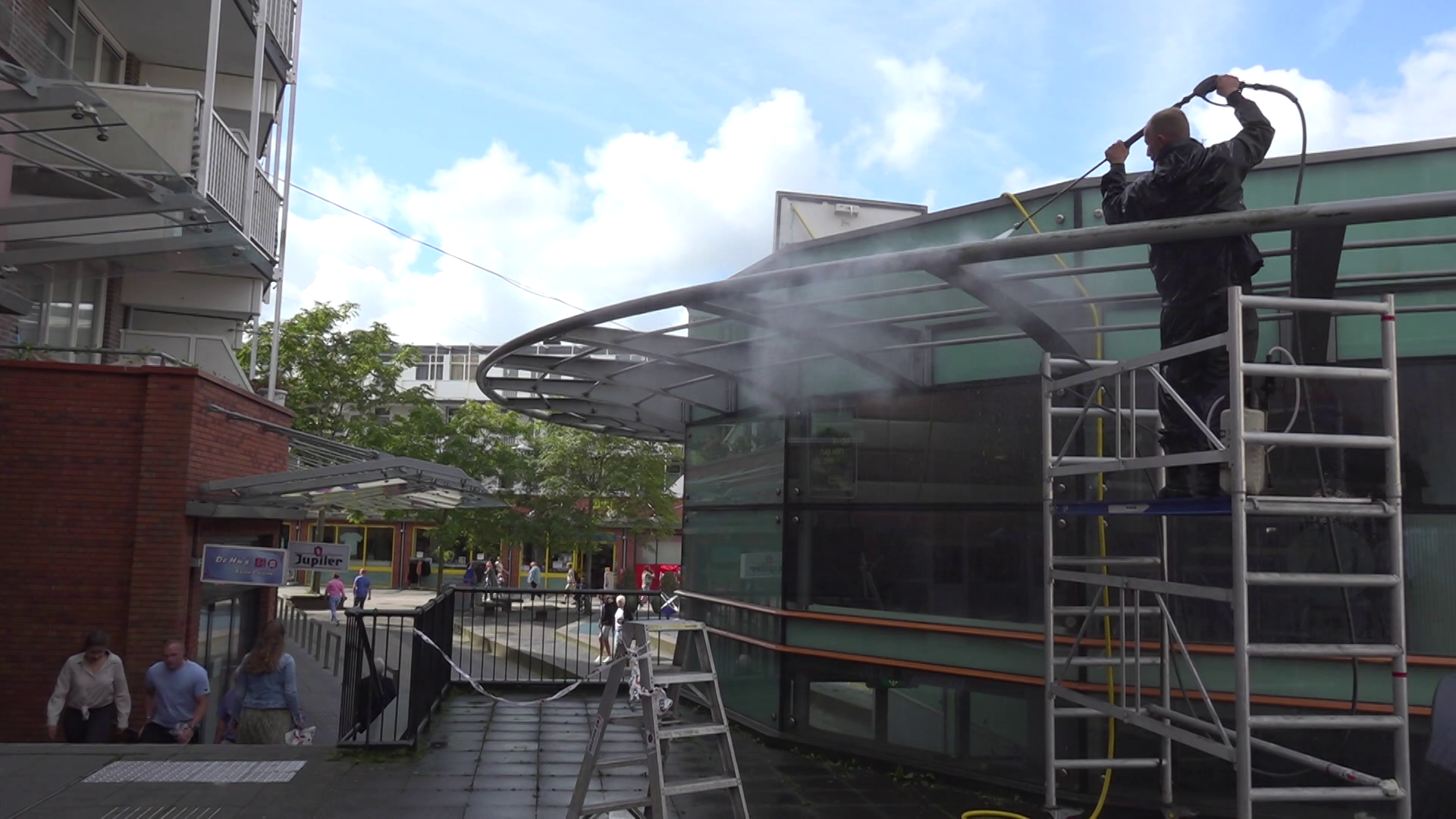 Man op steiger reinigt glazen overkapping met hogedrukspuit bij gebouw, met mensen die beneden lopen in een stedelijke omgeving.