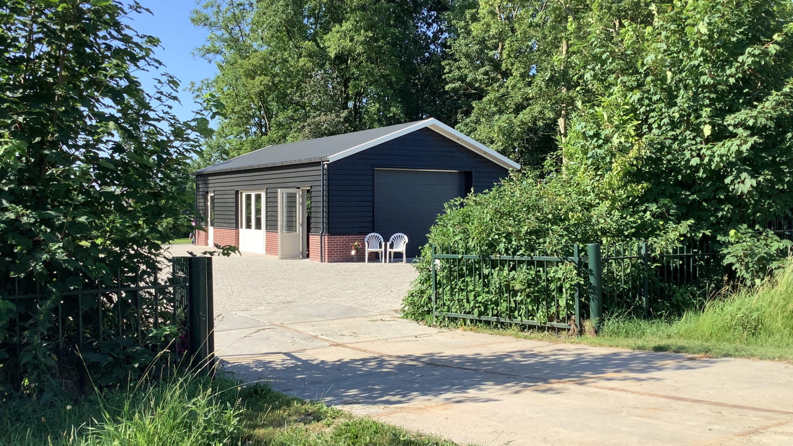 Schuur met zwart houten gevelbekleding en een grijs dak, gelegen in een groene omgeving met bomen en struiken, twee witte tuinstoelen voor de schuur.