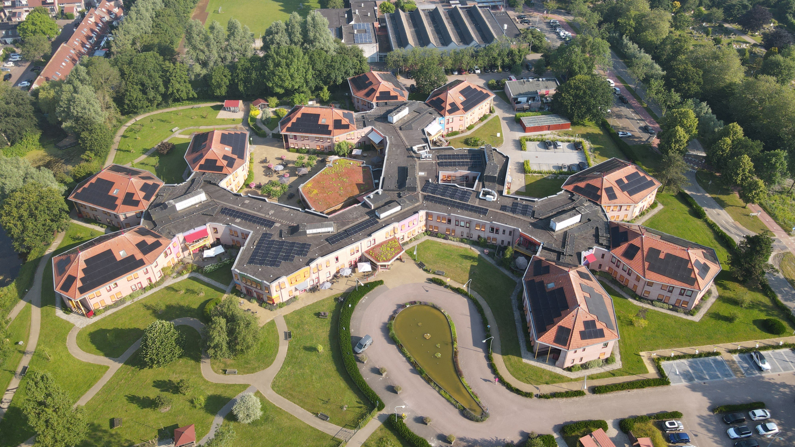 Luchtfoto van een groot, stervormig gebouw omringd door groen, lanen en parkeerplaatsen; het dak is voorzien van zonnepanelen.