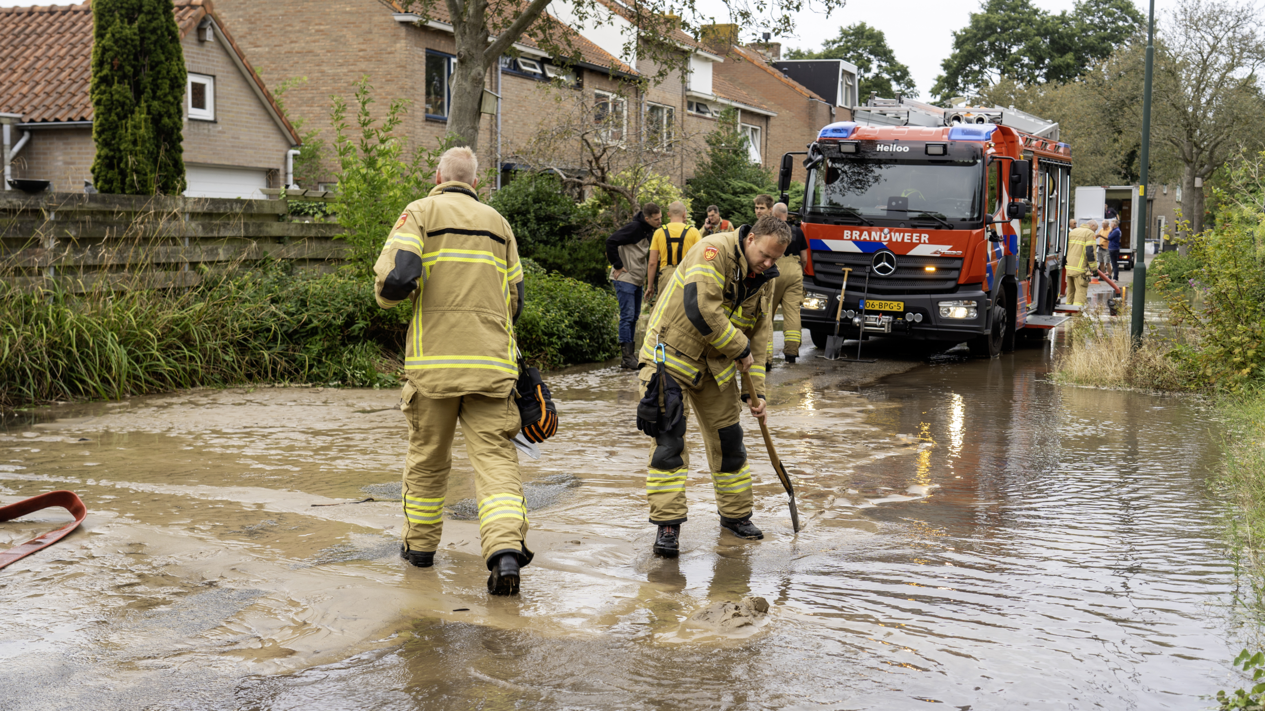 Brandweerlieden werken op een met modder overstroomde straat bij een brandweerwagen in een woonwijk.