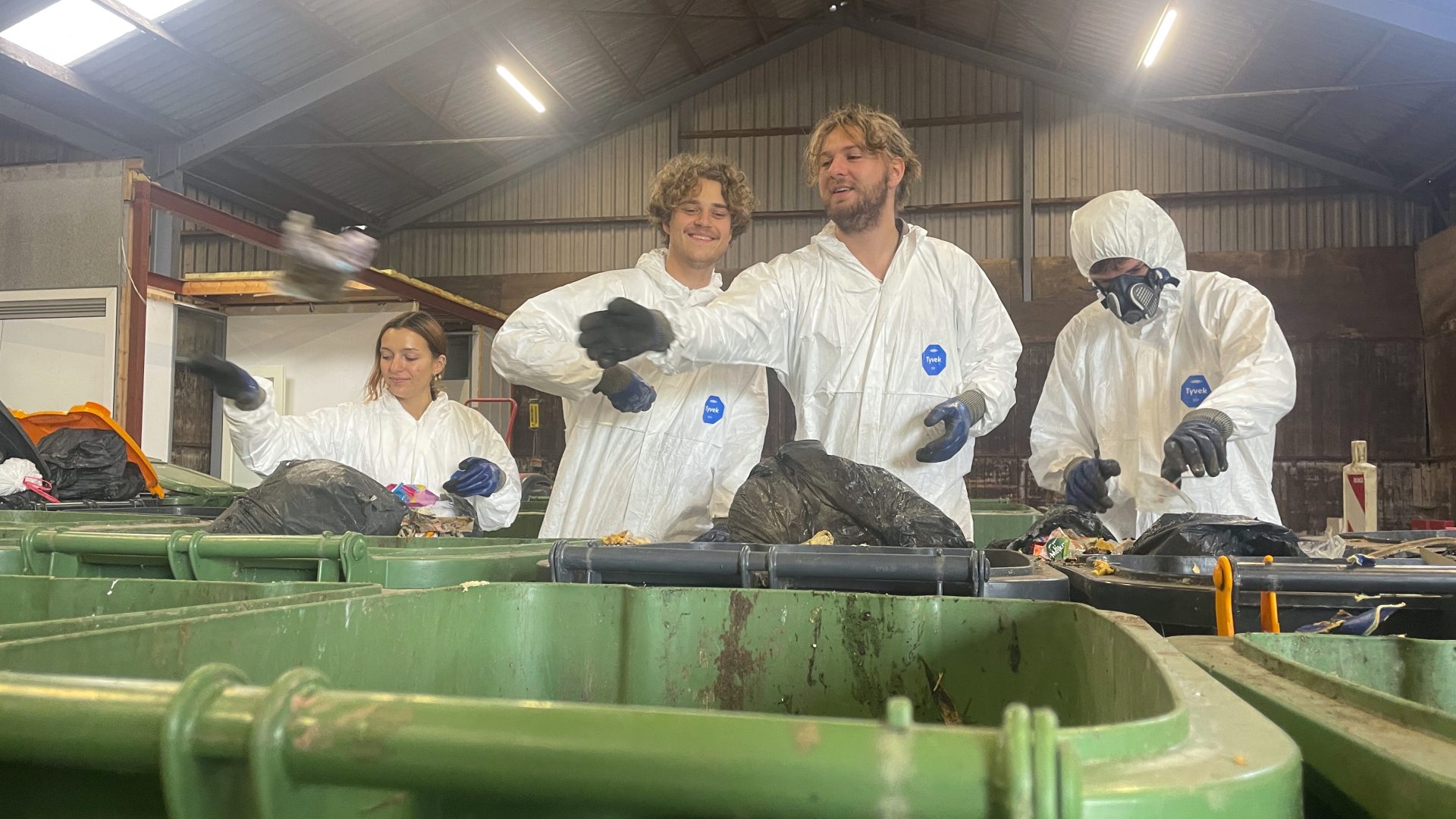 Vier mensen in witte beschermende pakken sorteren afval in groene containers in een loods.