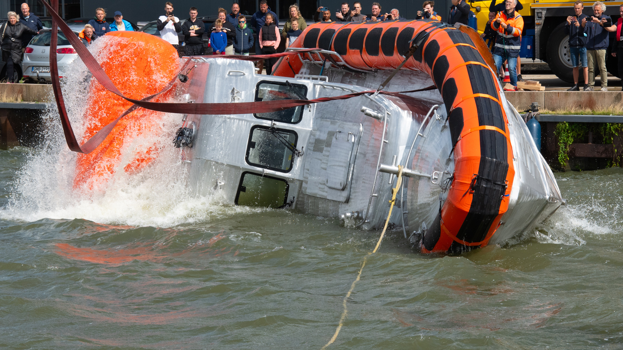 Mensen kijken toe hoe een grote metalen capsule met opblaasbaar oranje drijfvermogen in het water wordt gelanceerd.