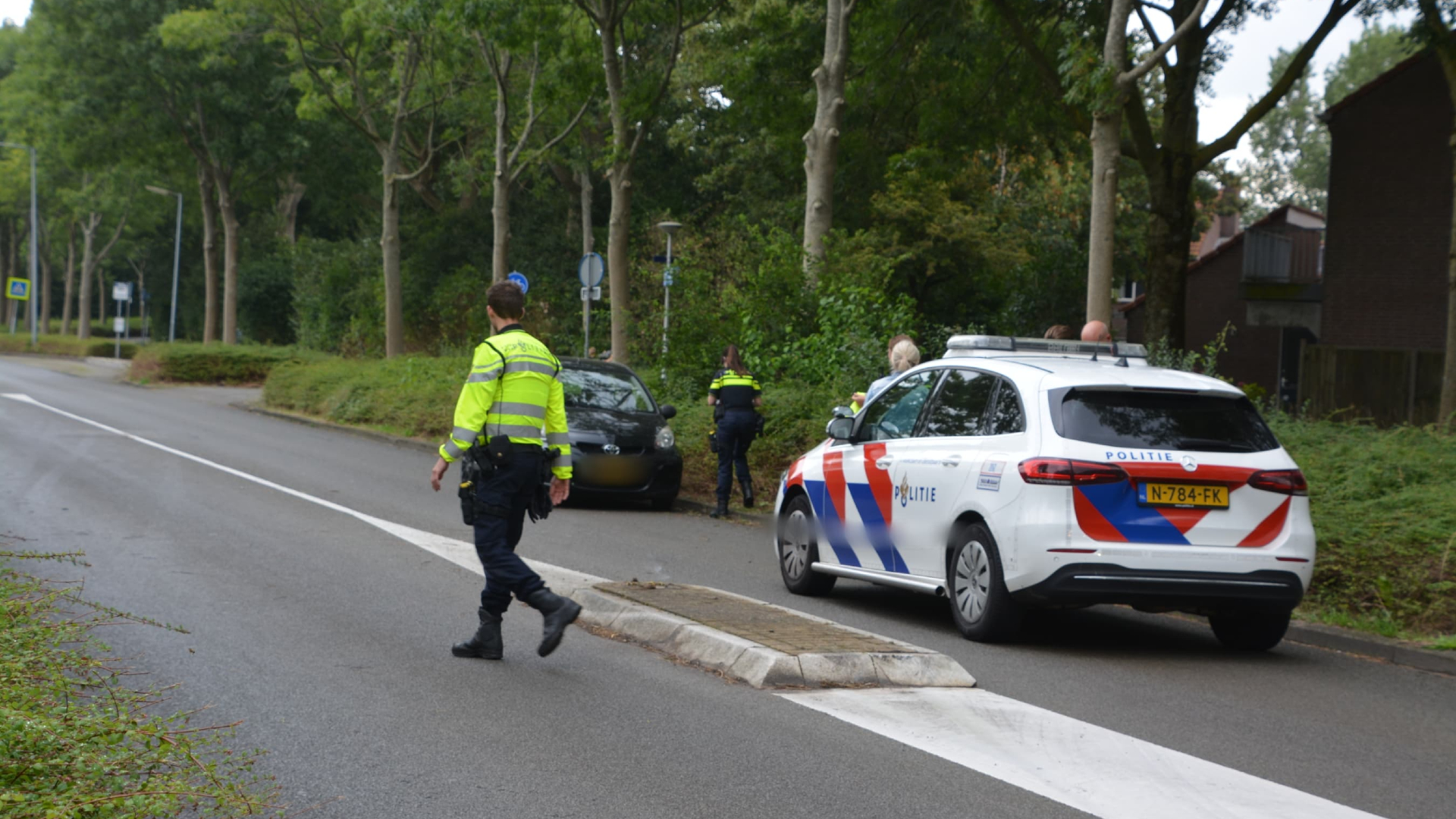 Politieagenten en een politiewagen bij een incident langs een weg met bomen.