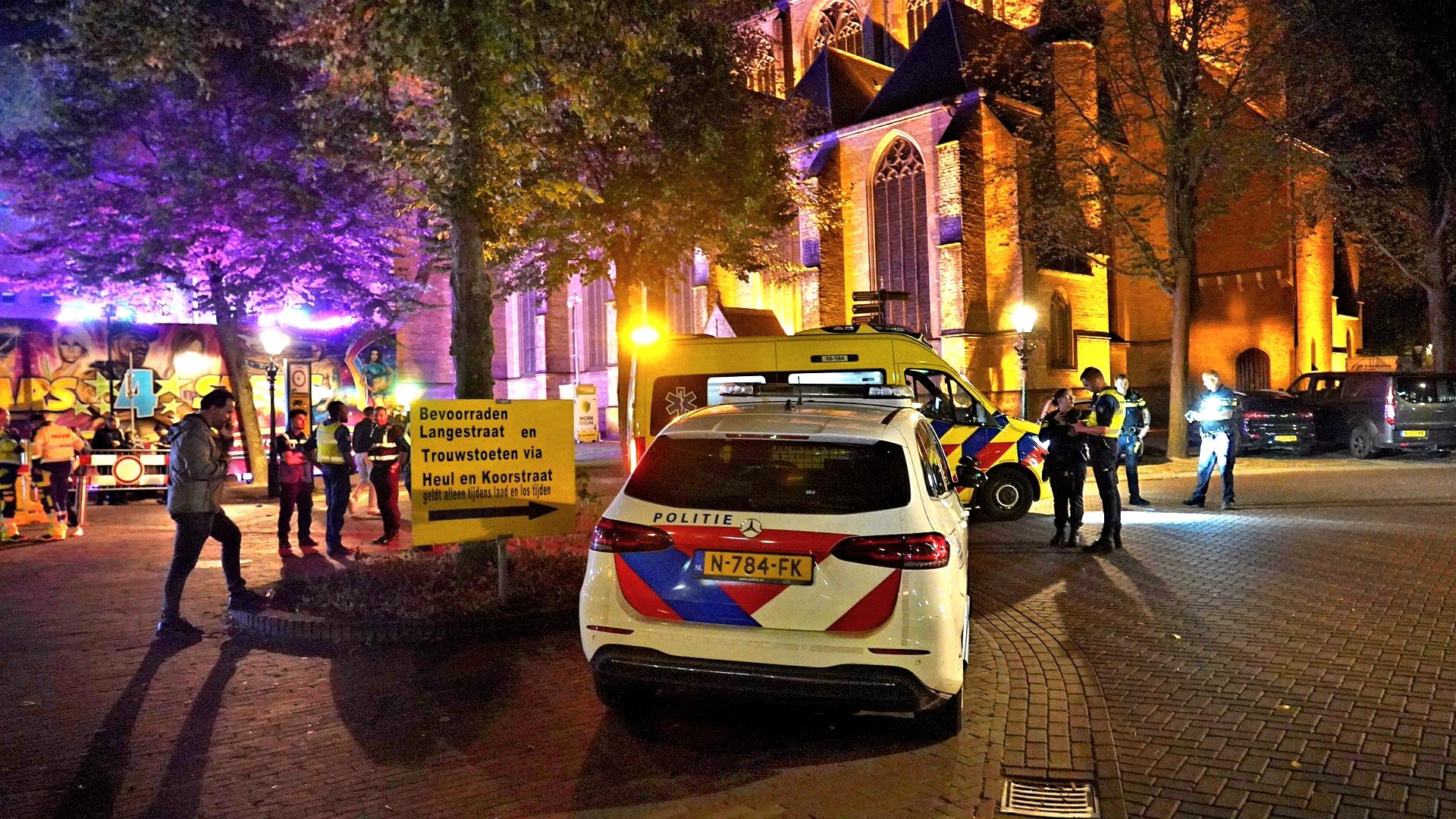 Politie- en ambulancevoertuigen bij een verlicht gebouw in de avond, met agenten en andere mensen in reflecterende kleding op de achtergrond.