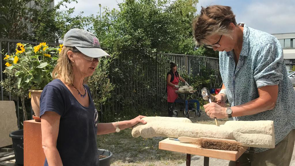 Twee vrouwen werken samen aan een houtbewerkingsproject buiten, terwijl een derde persoon op de achtergrond bezig is.