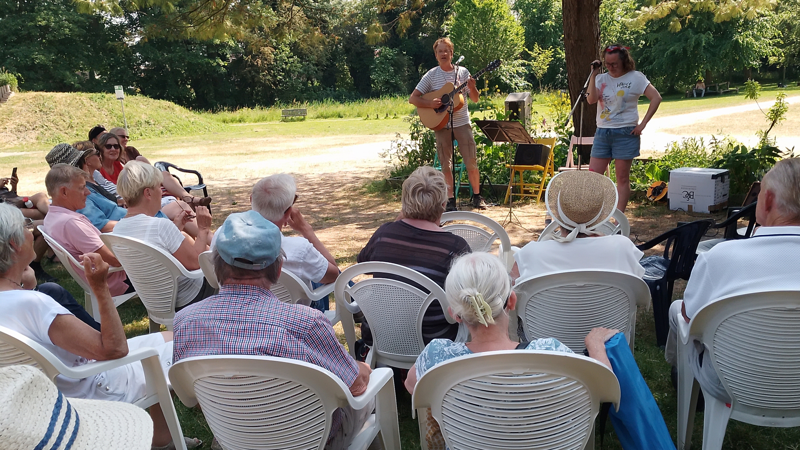 Mensen kijken naar een live muziekoptreden in een park terwijl ze op witte stoelen zitten.