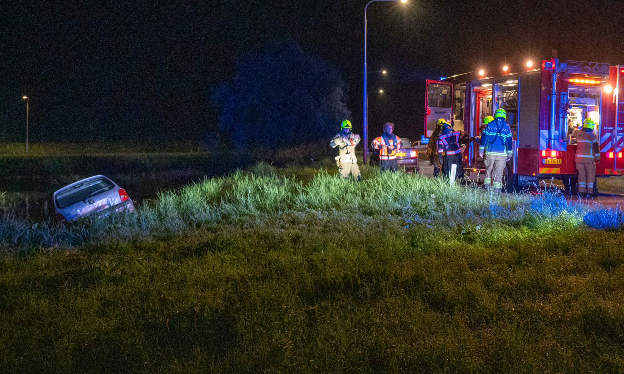 Brandweerlieden bij een auto-ongeluk langs een donkere weg, waarbij een auto in een greppel is beland.