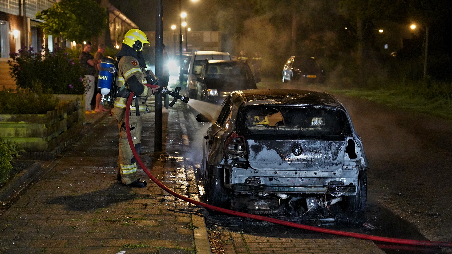 Brandweerman blust in de nacht een uitgebrande auto op een woonstraat.