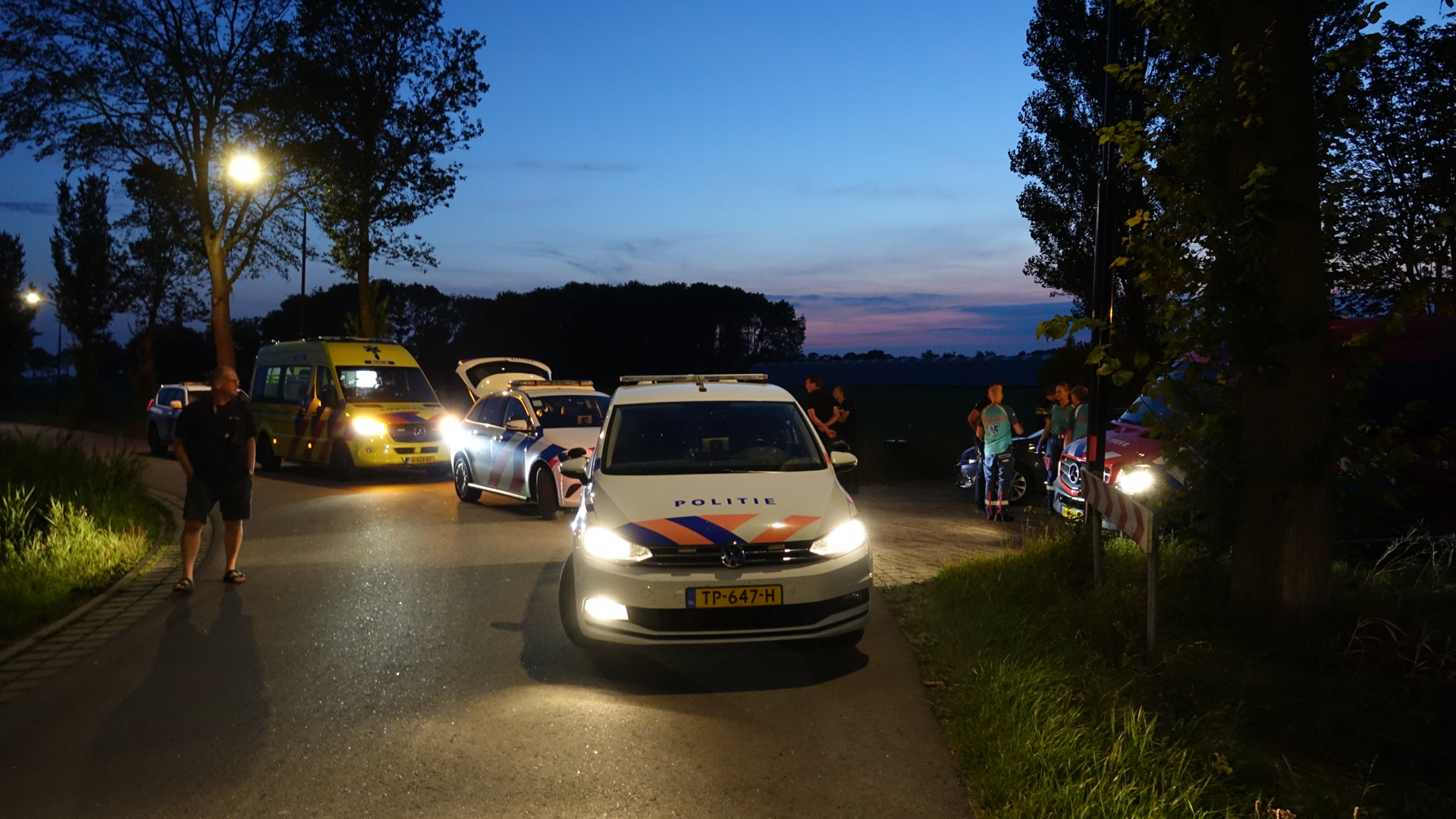 Politie- en ambulancescene op een weg bij zonsondergang, met lichten van de voertuigen aan en mensen die buiten staan en praten.