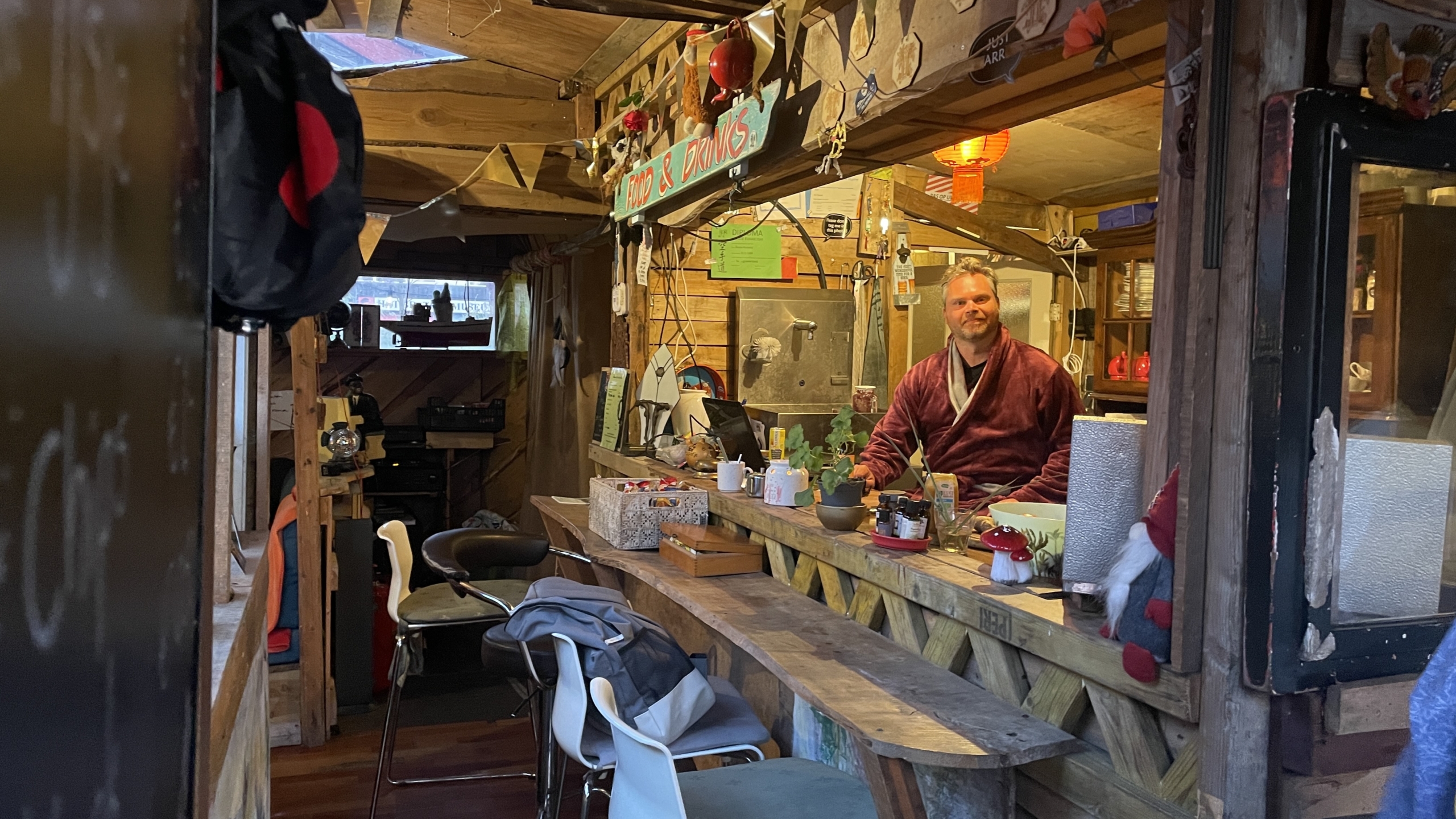 Man in een houten bar met een bord "Food & Drinks", omgeven door decoraties en planten.